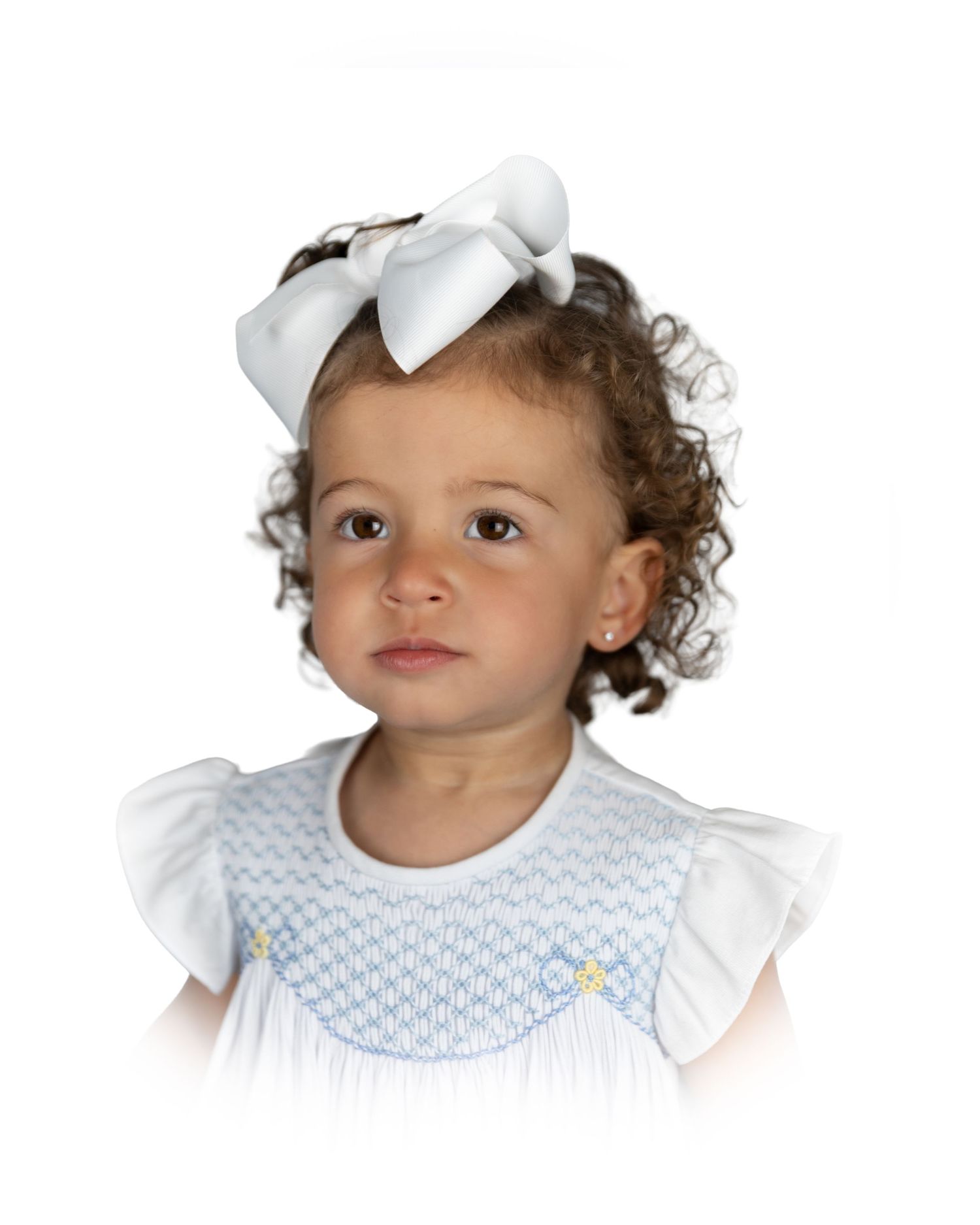 Toddler girl with curly hair wearing a white smocked dress with a white bow headband.
