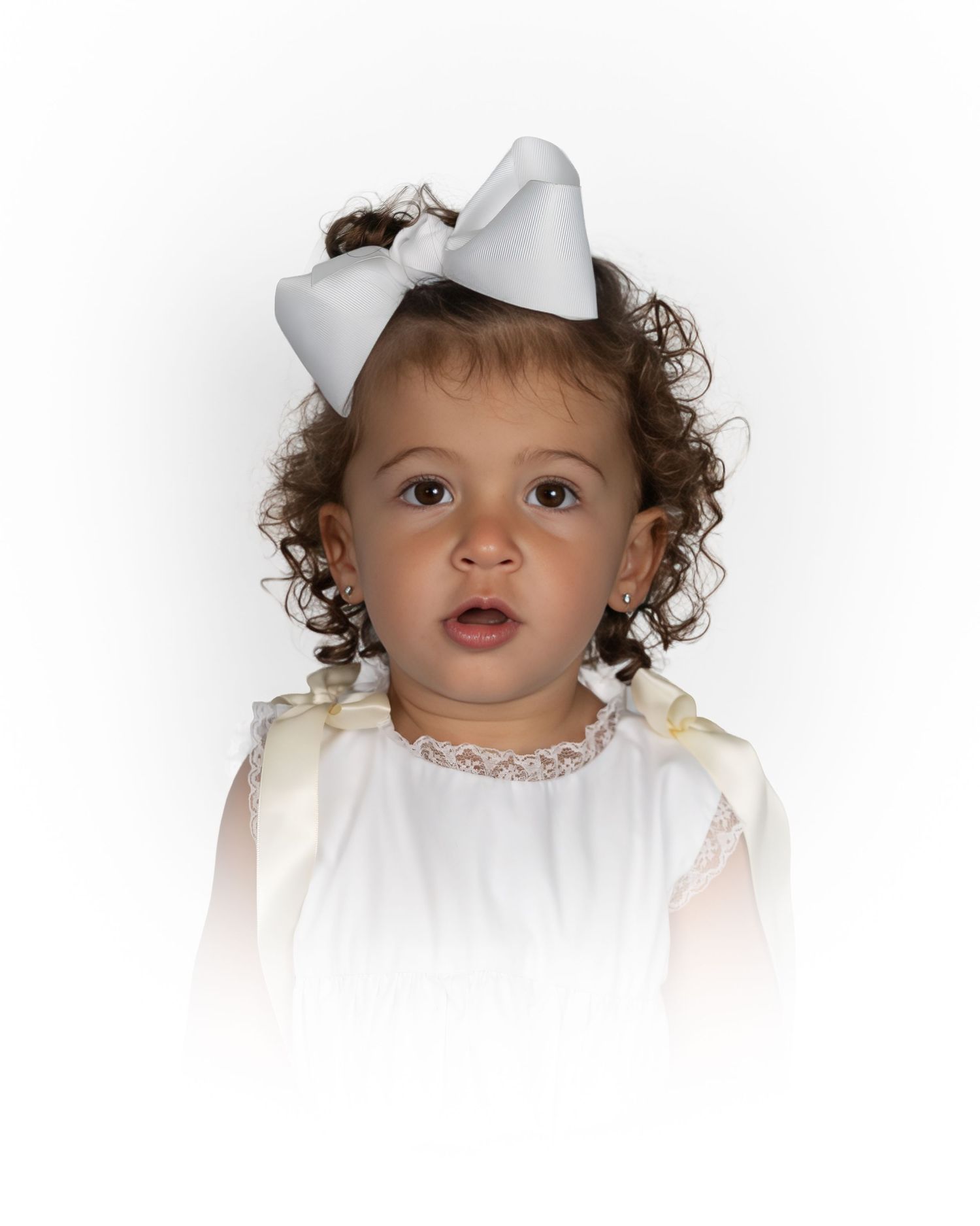 Baby girl with curly brown hair wearing a white bow and white lace dress.
