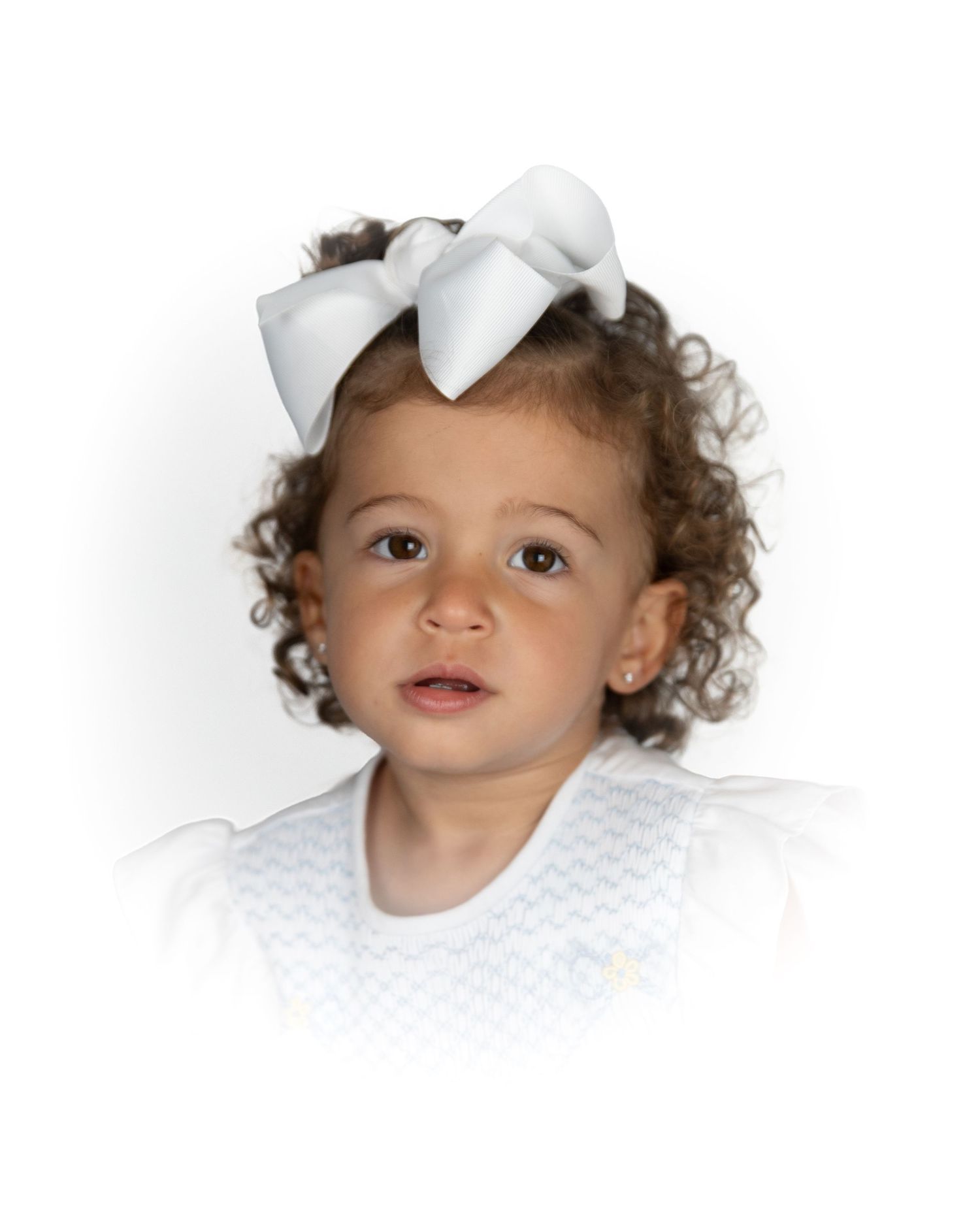 Toddler girl with curly hair wearing a large white bow and white outfit.