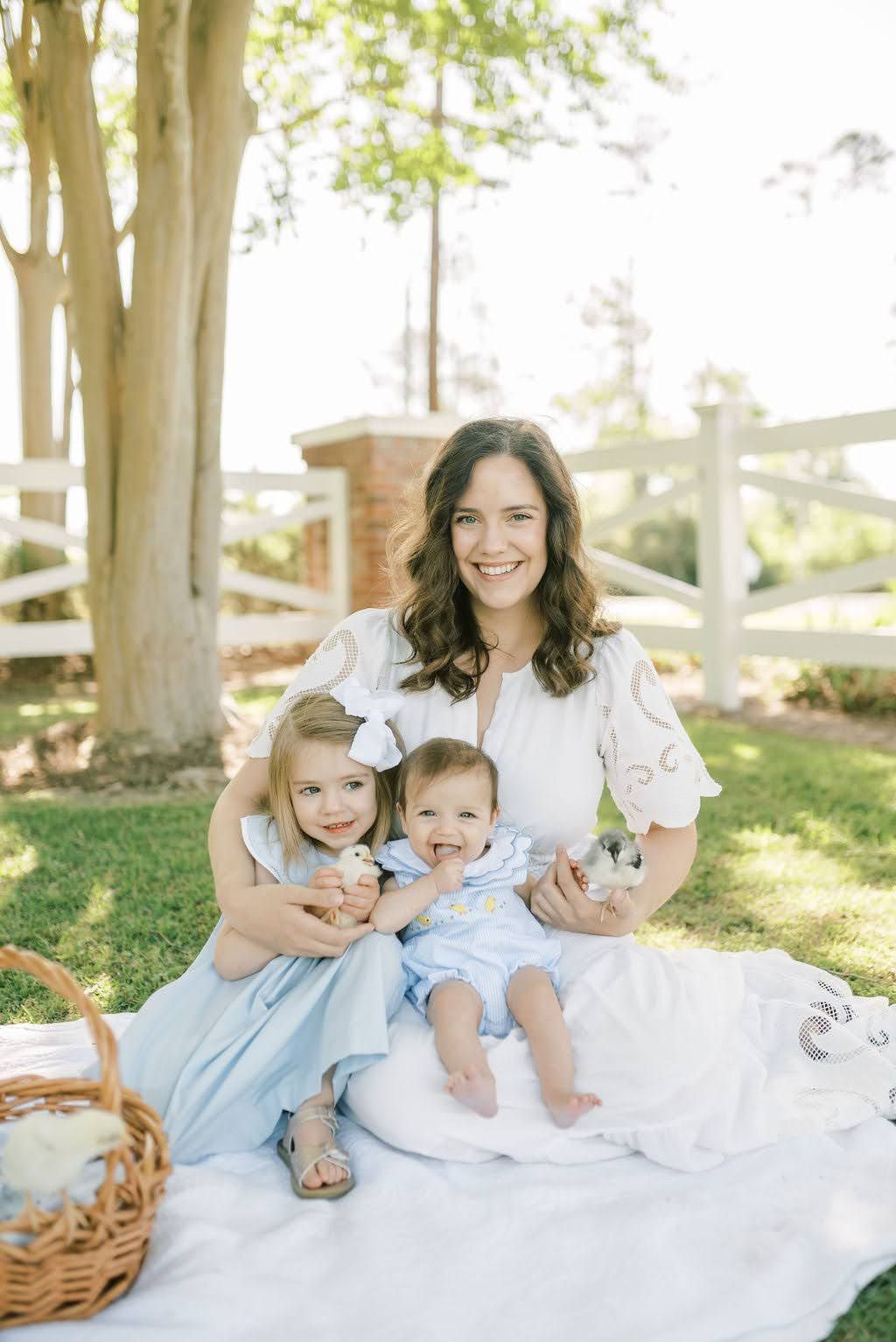 A smiling mother sits on a blanket outdoors with her young daughter and baby in matching outfits.