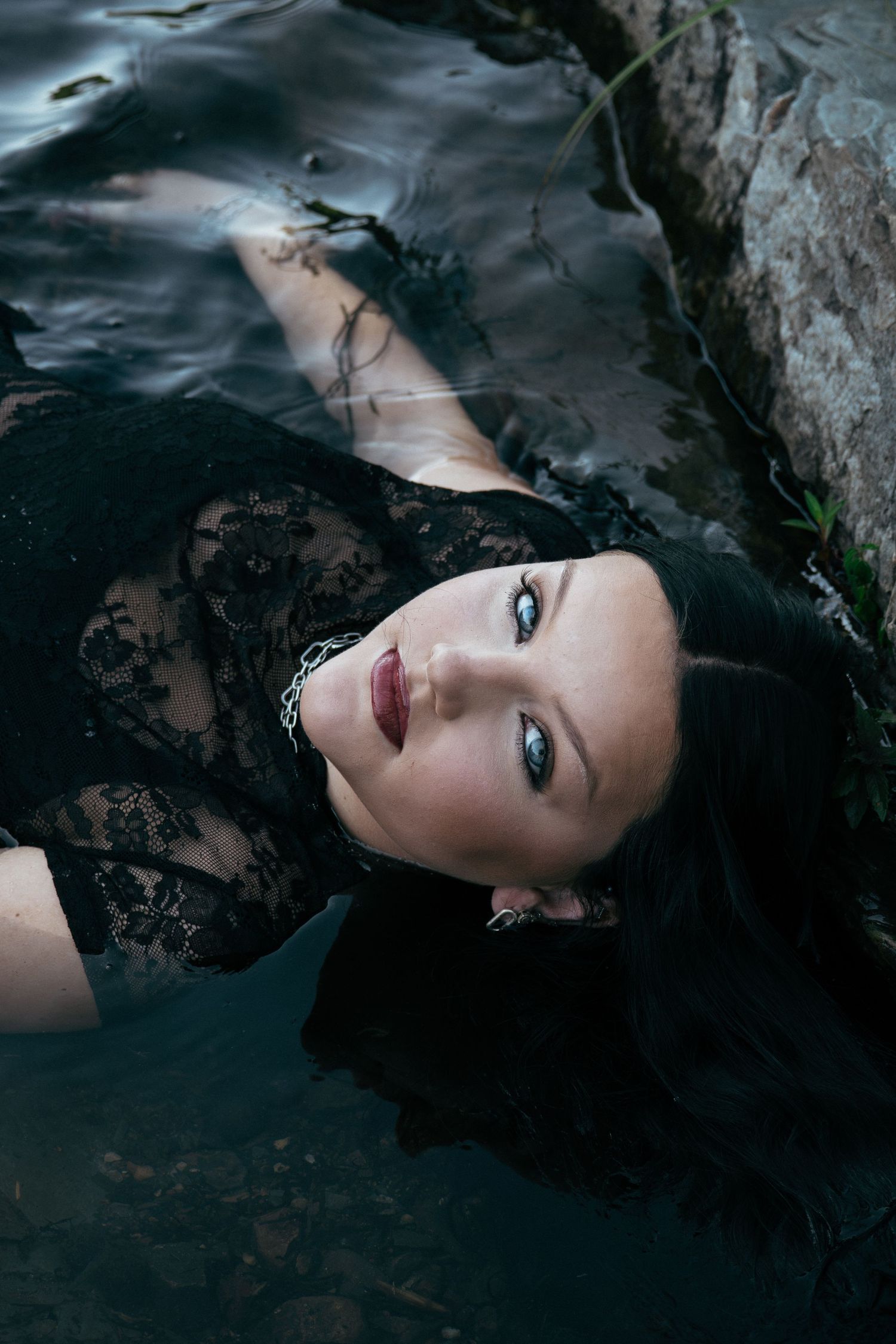 Woman in black lace dress lying in dark water, gothic aesthetic portrait with blue eyes and dark hair.