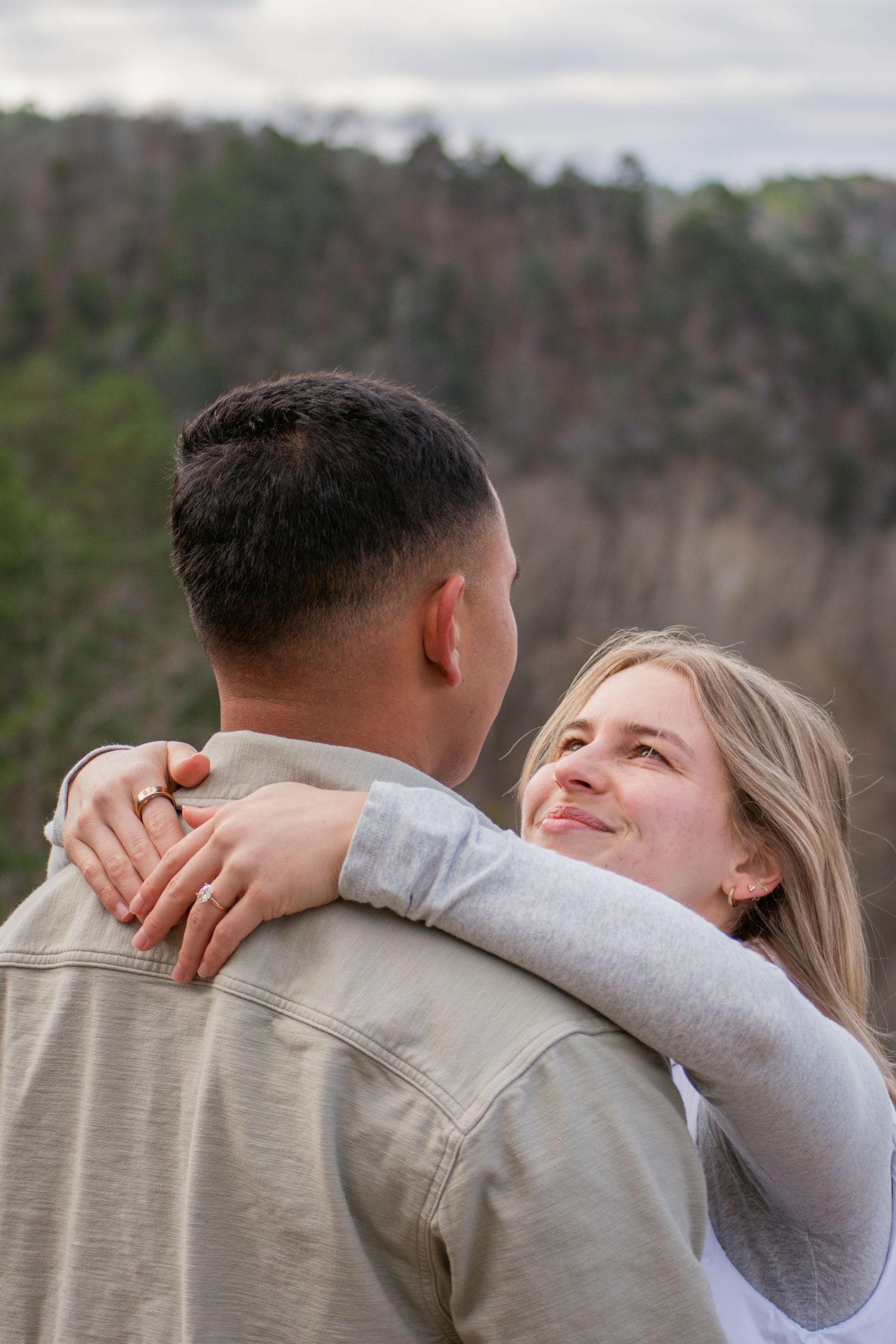 Smiling woman with blonde hair embracing a man outdoors with lush green hills in the background.