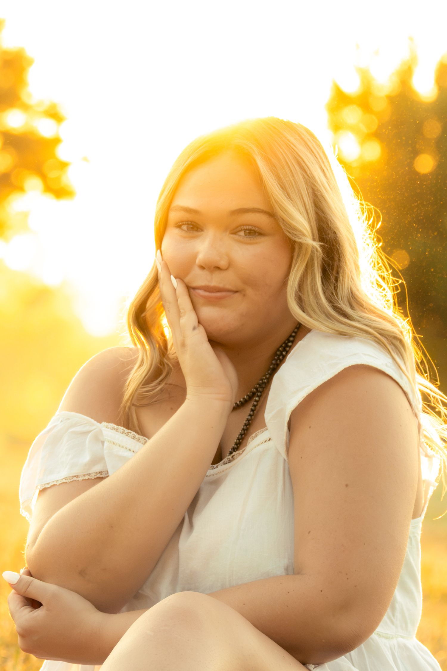 Young blonde woman in white dress poses outdoors in golden sunset light, hand on cheek, wearing beaded necklace.