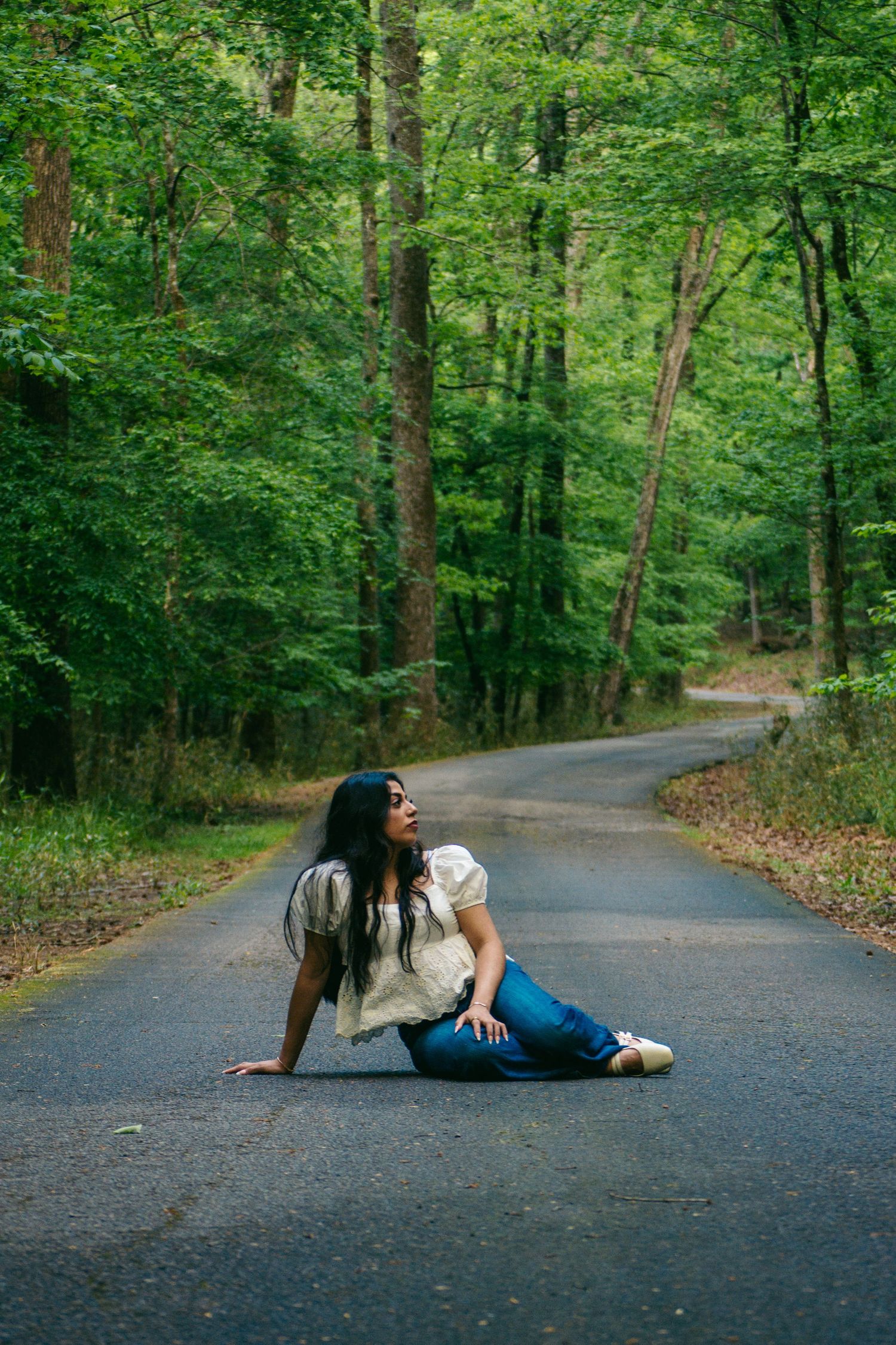 A young woman with long dark hair sits on a winding forest road surrounded by lush green trees.