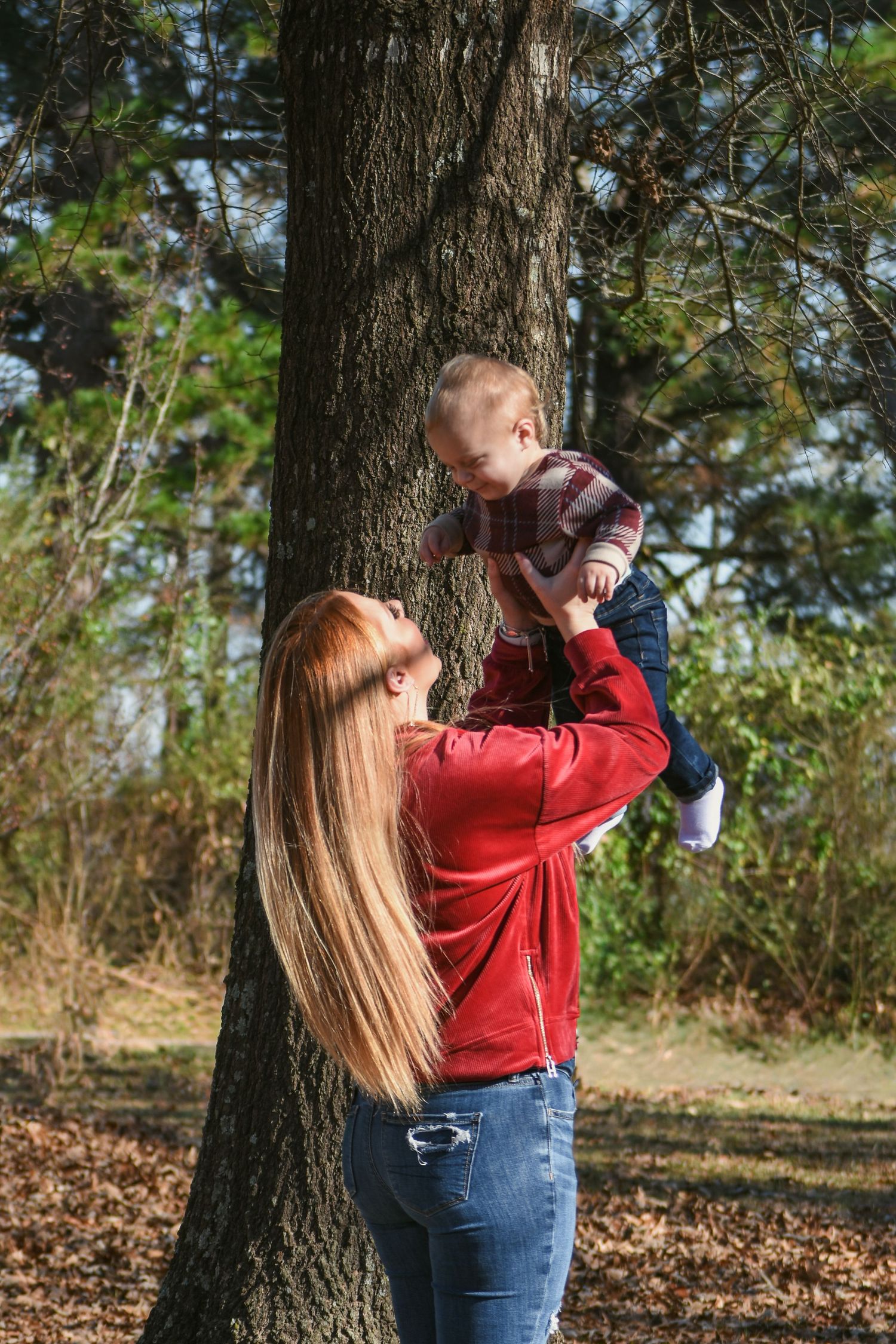 A red-haired mother lifts her smiling baby boy up in the air beside a large tree outdoors.