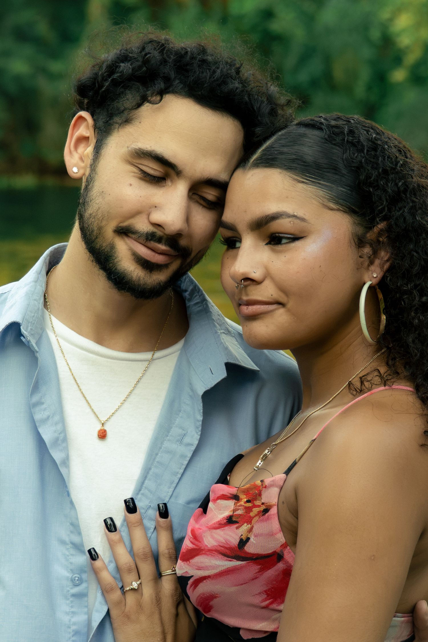 A young couple in a tender moment, the man in a blue shirt and woman in a floral top, touching foreheads outdoors.