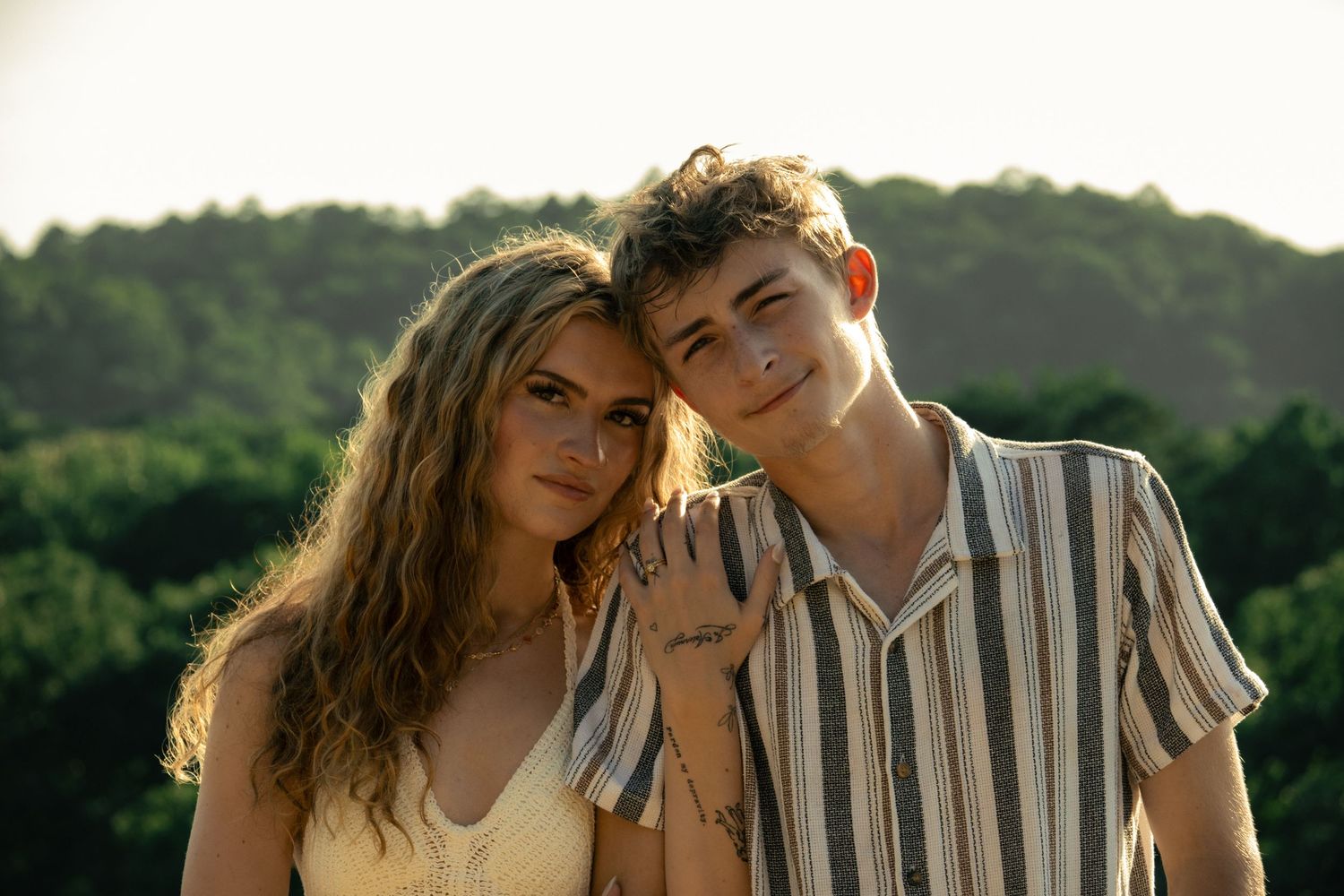 Young couple smiling together outdoors with lush green trees in the background on a sunny day.
