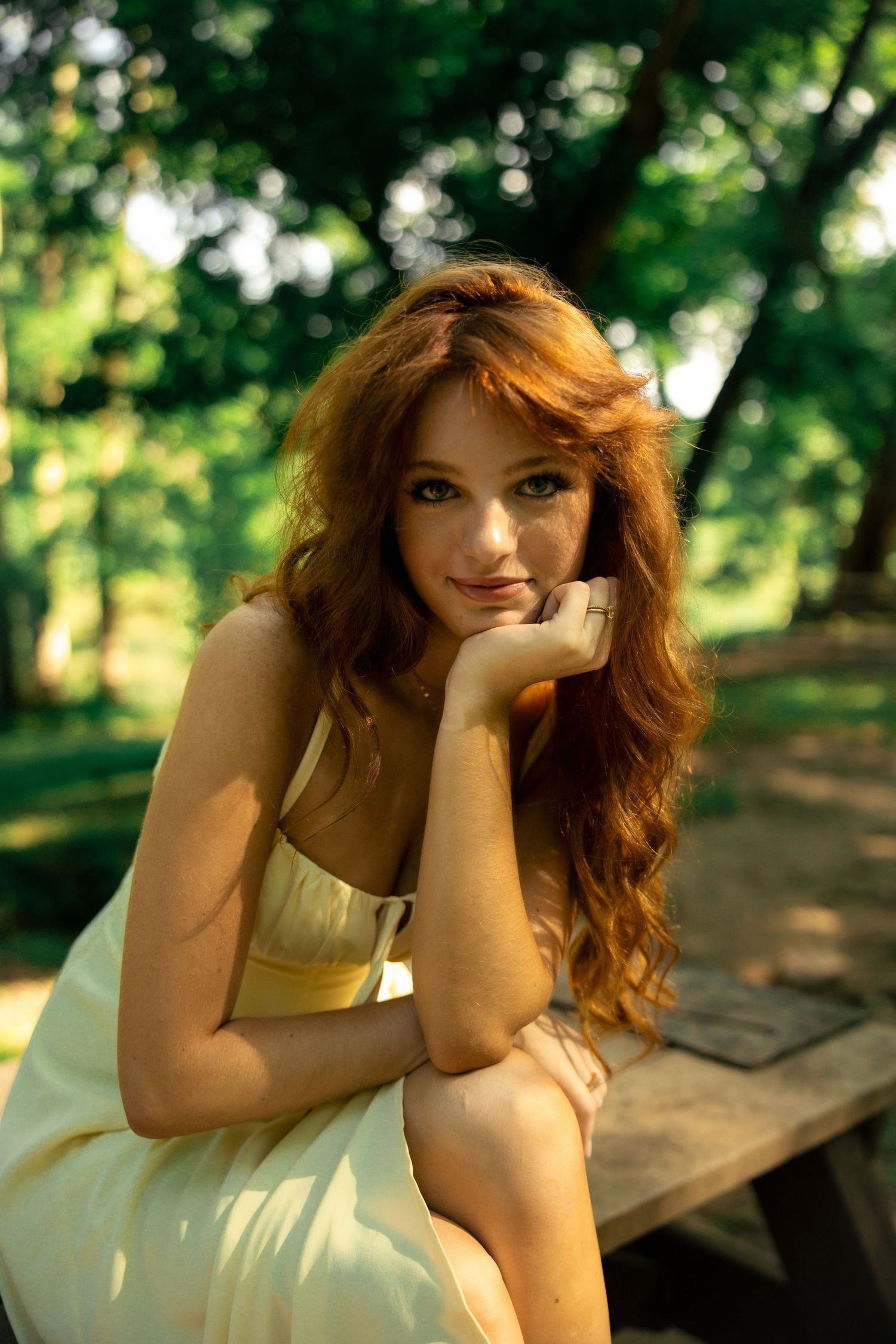 Young woman with long red wavy hair in a yellow dress sitting at a wooden picnic table in a lush green park.
