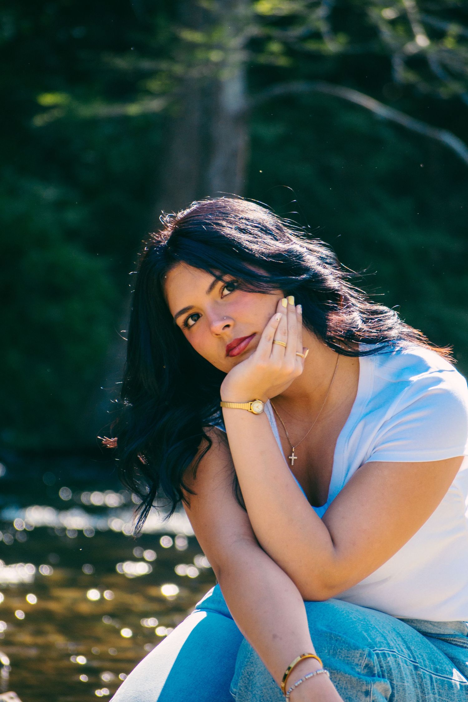 Young woman with dark wavy hair poses thoughtfully by a shimmering forest stream in white top and jeans.