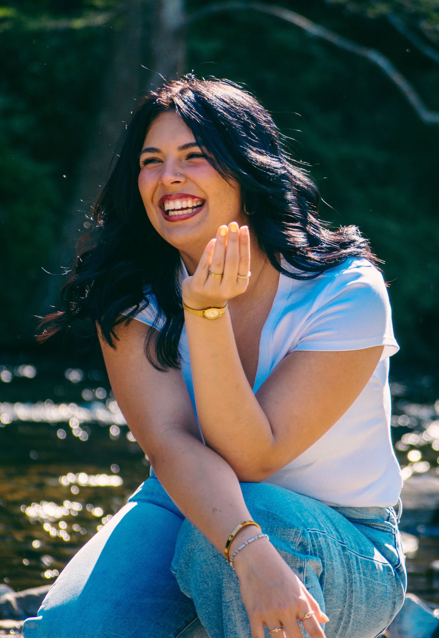 Young woman with dark wavy hair laughs joyfully while sitting near a sunlit creek wearing a white t-shirt and jeans.