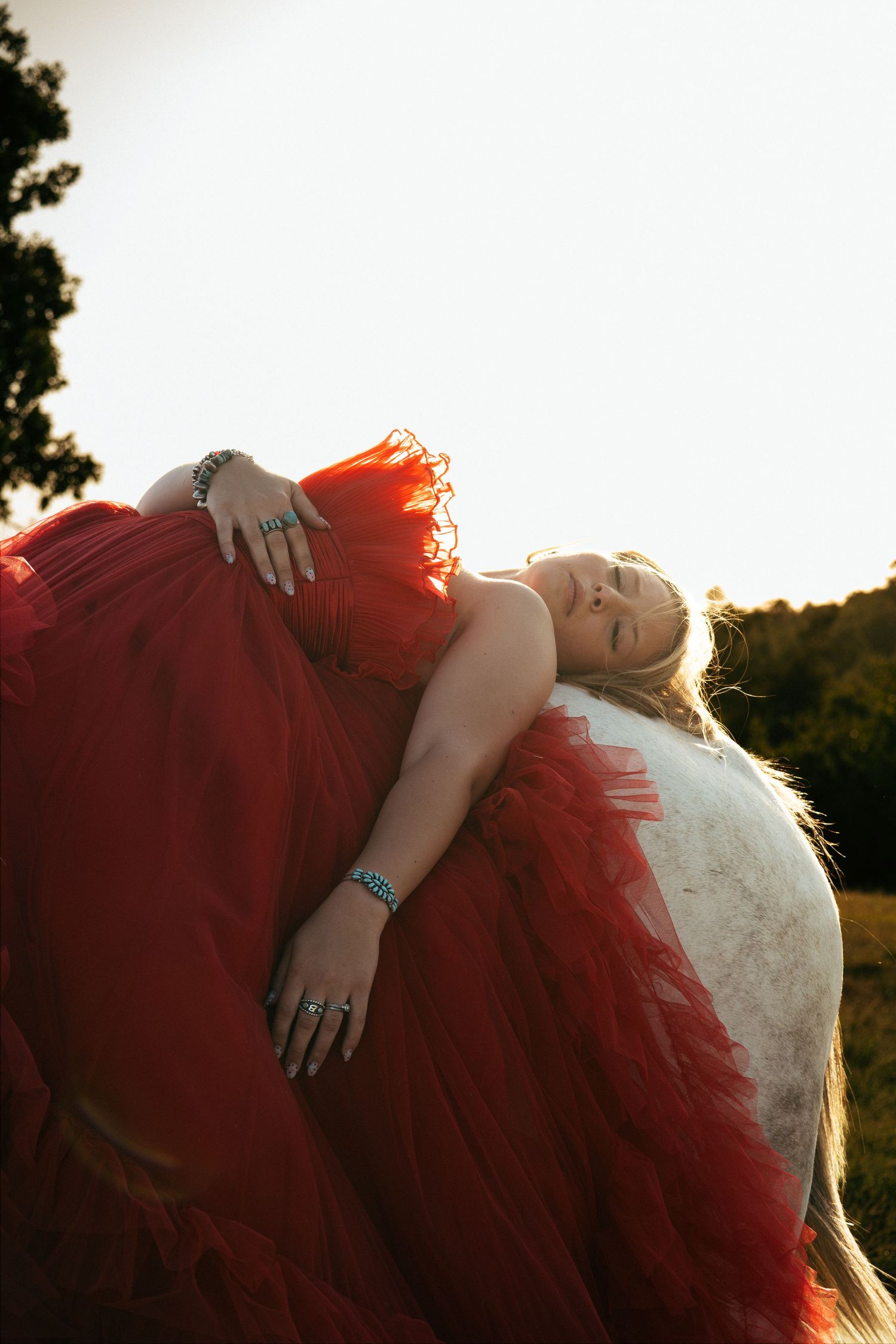Woman in red tulle gown leaning on white horse at golden hour in a field, wearing turquoise jewelry.