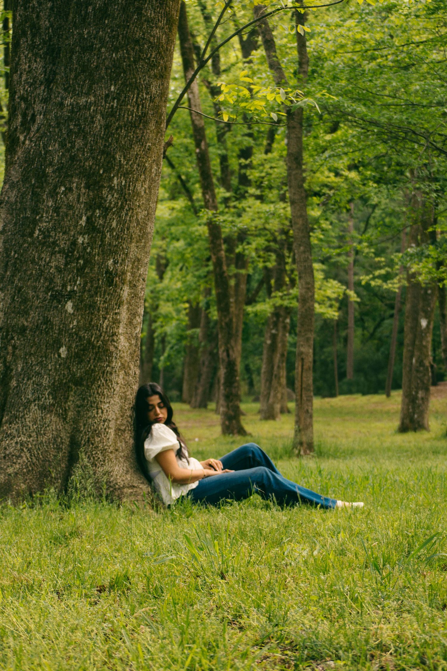 Young woman in white top and jeans relaxes against a large tree trunk in a lush green sunlit forest.