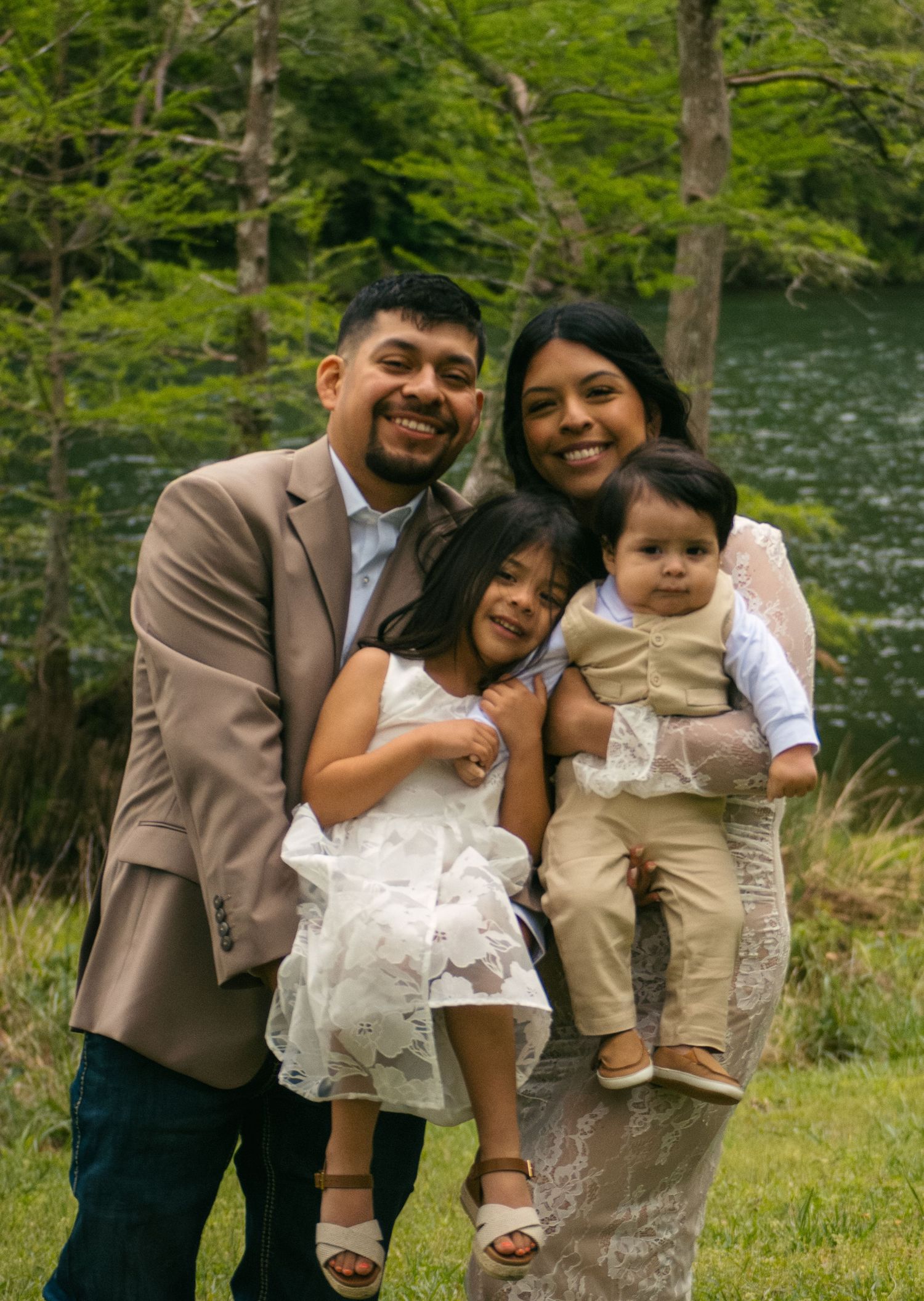 Happy Hispanic family of four smiling together outdoors near a green tree-lined river.