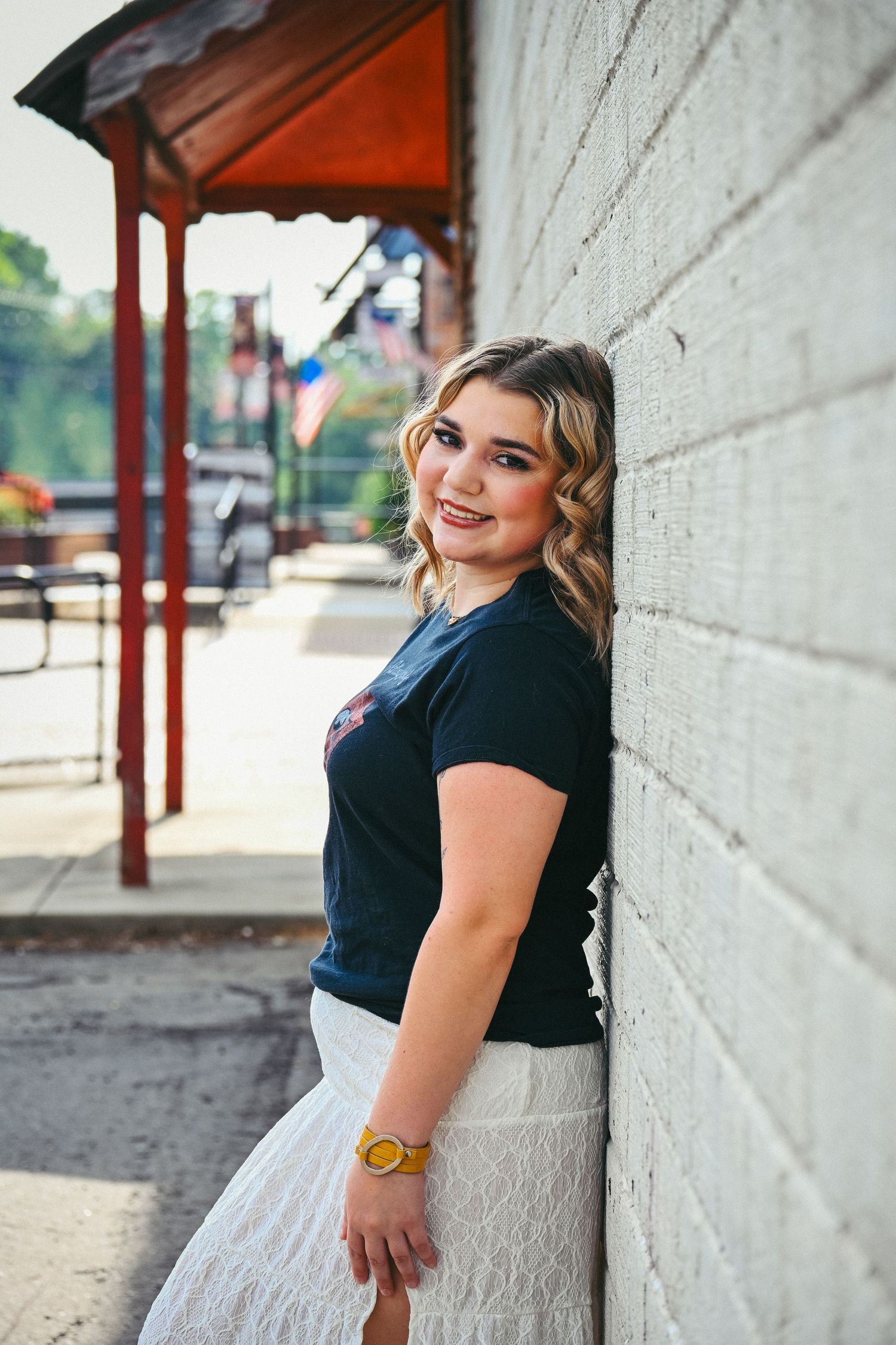Smiling blonde teen girl in navy tee and white lace skirt leaning against a brick wall on a sunny street.