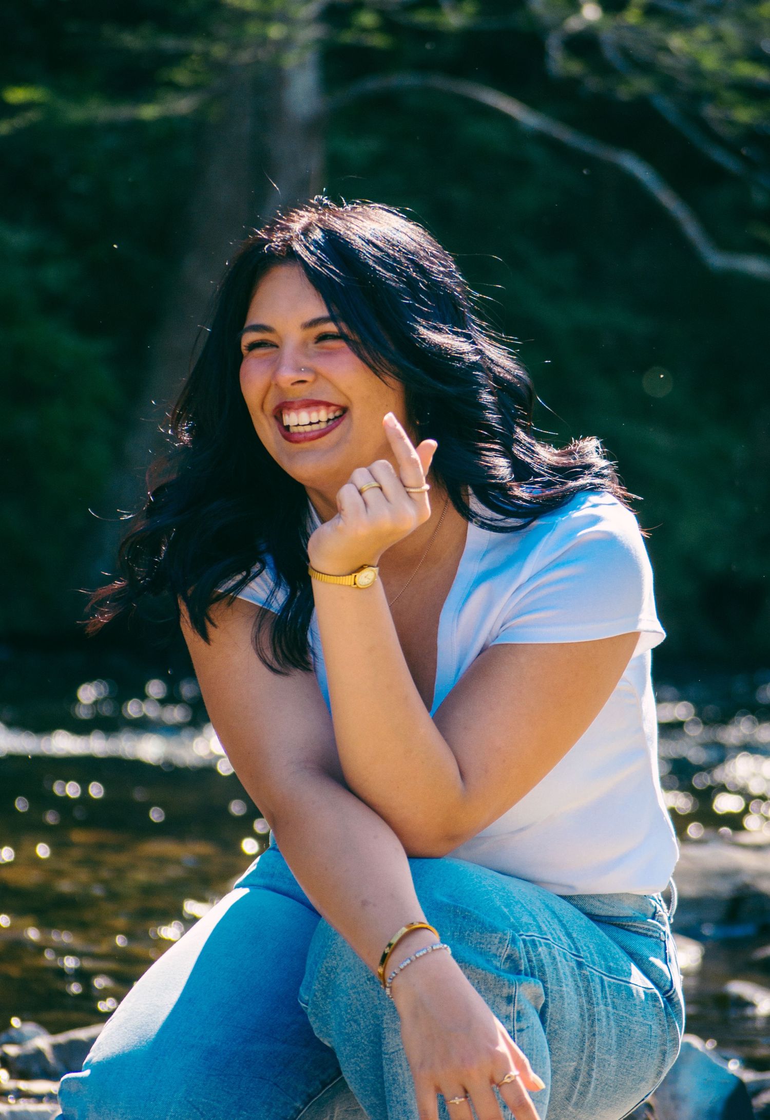Joyful young woman with dark wavy hair laughs while sitting by a sunlit forest stream in white tee.