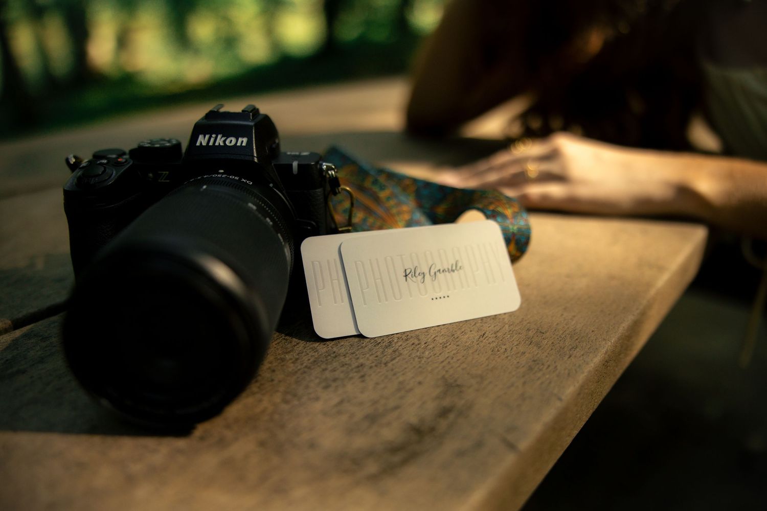 Nikon camera and business card on wooden table with blurred woman in background in outdoor setting.