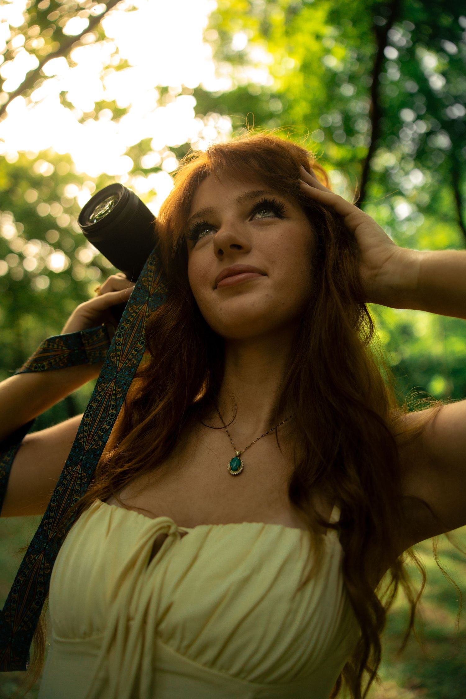 Red-haired woman in white dress holding a sword scabbard in a sunlit forest, wearing a green gemstone necklace.