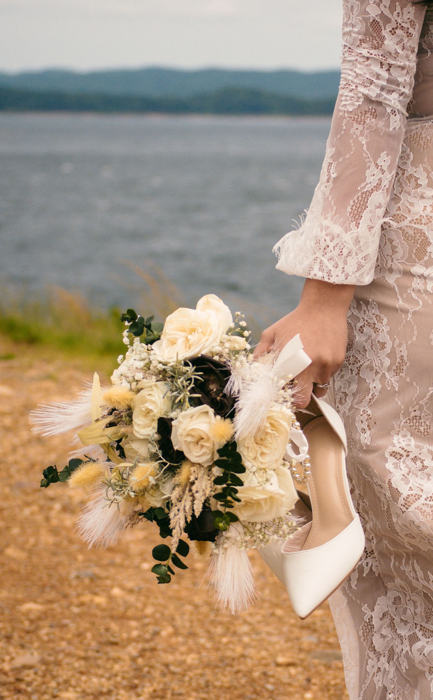 Bride in lace wedding dress holding white rose bouquet and heels near scenic waterfront.