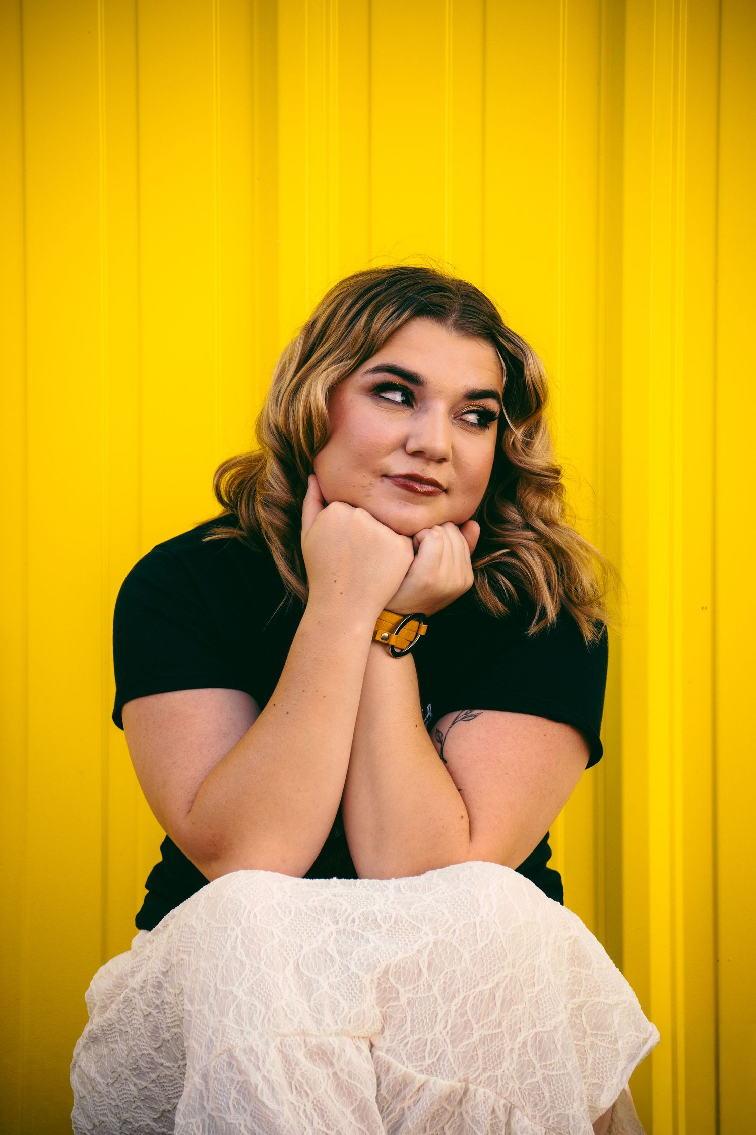 Young woman with wavy blonde hair rests her chin on her hands against a bold yellow background wearing a black top.