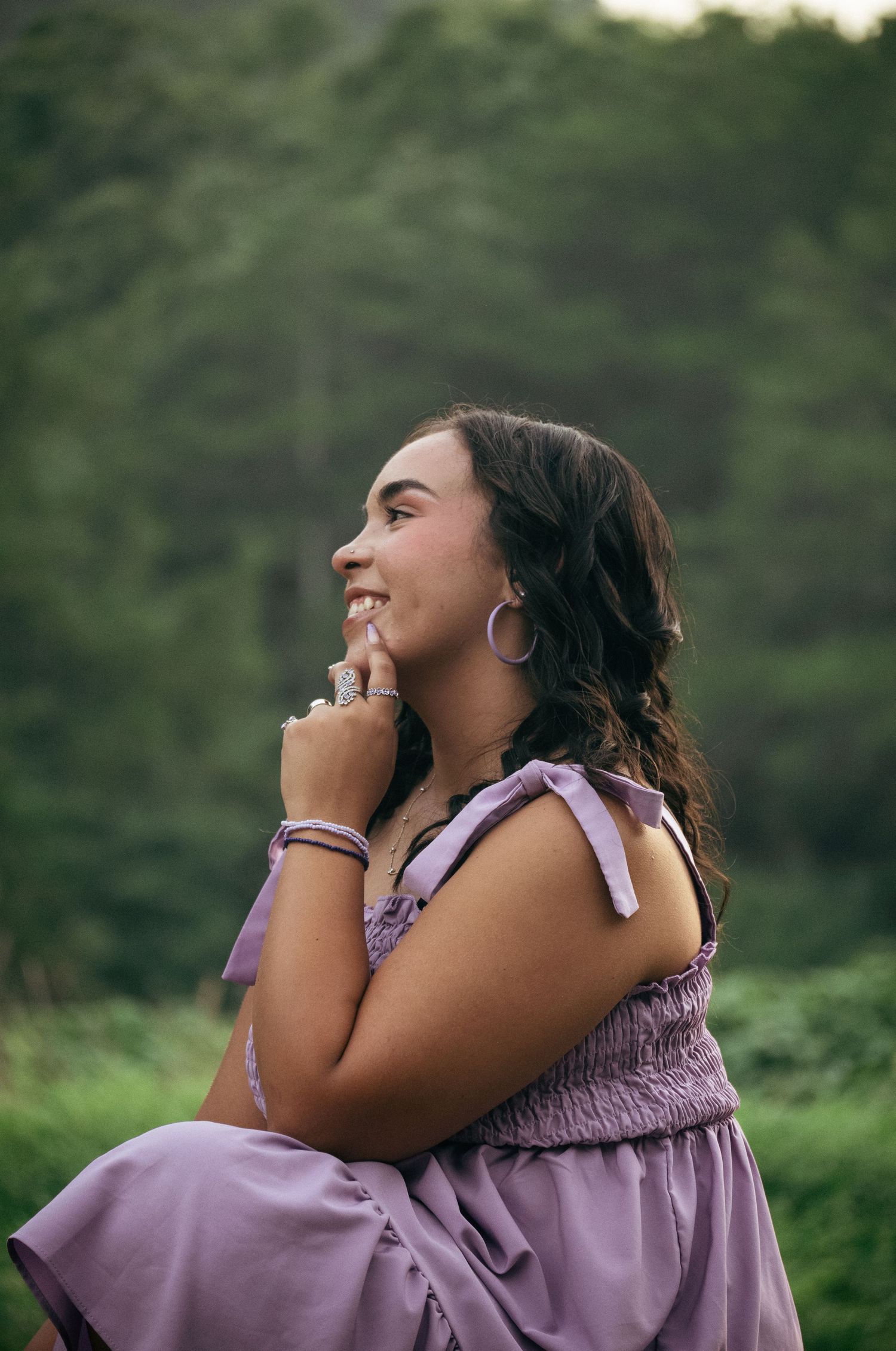 Young woman in lavender dress smiling outdoors, wearing hoop earrings and rings, with green trees in background.