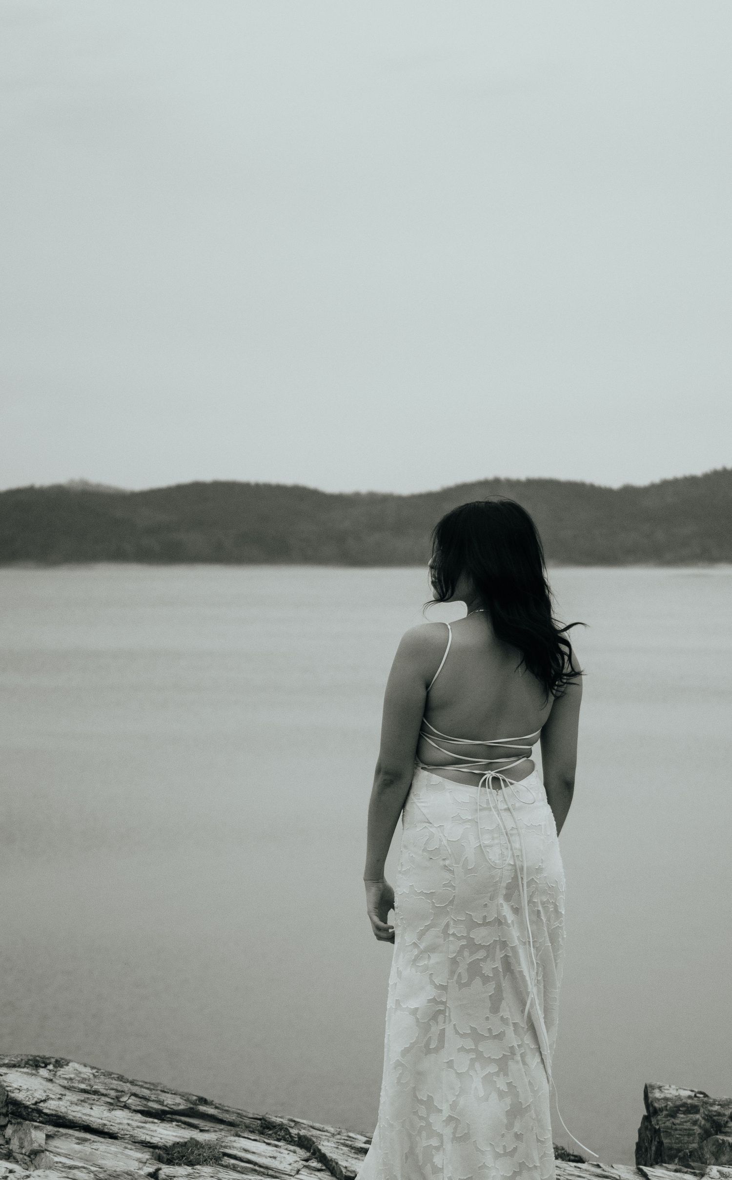 Woman in elegant white lace backless gown stands on rocky shore gazing at calm lake and distant hills.