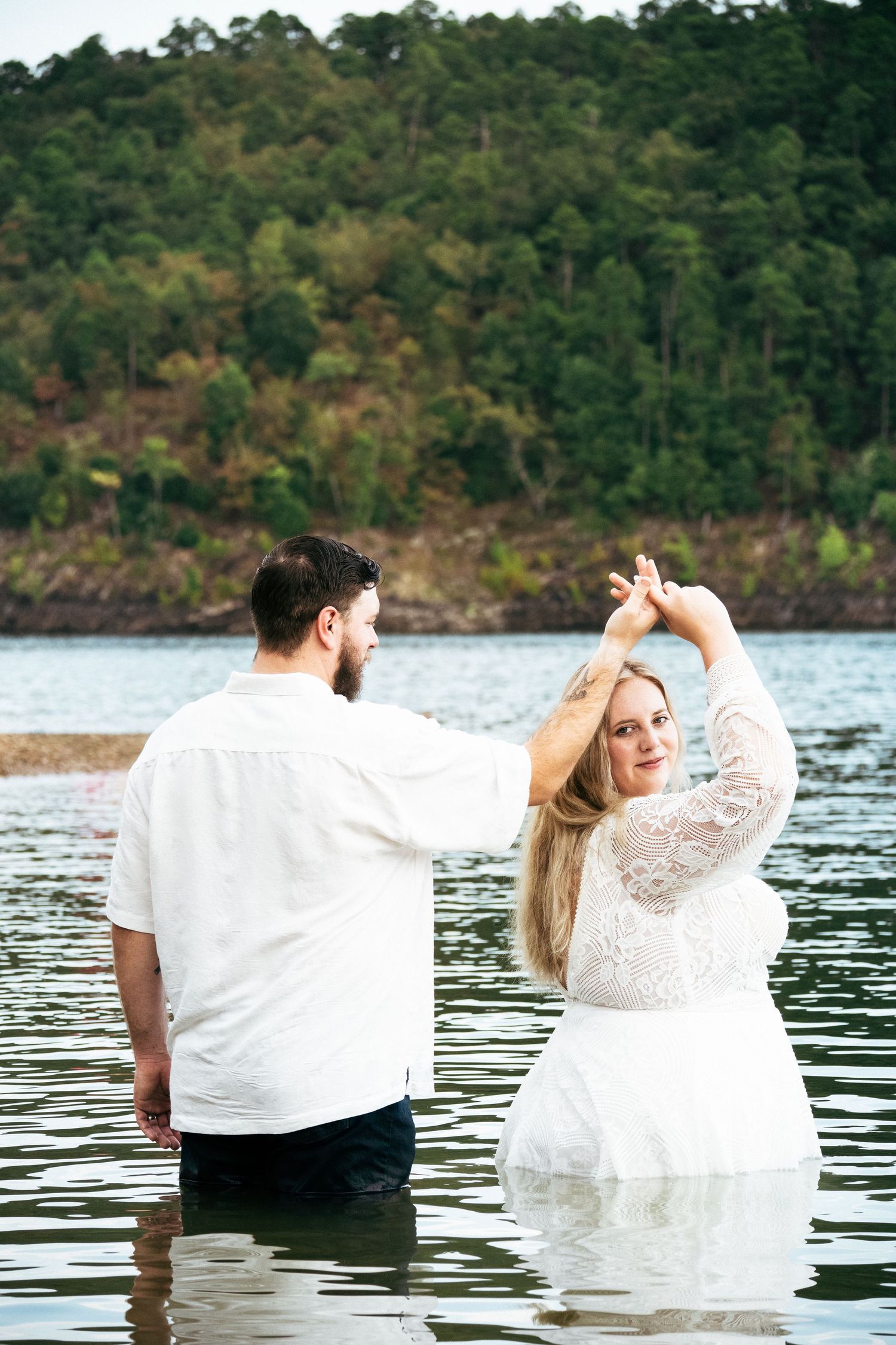 Couple in white attire standing in a lake, woman raising arms gracefully with lush green hills in background.
