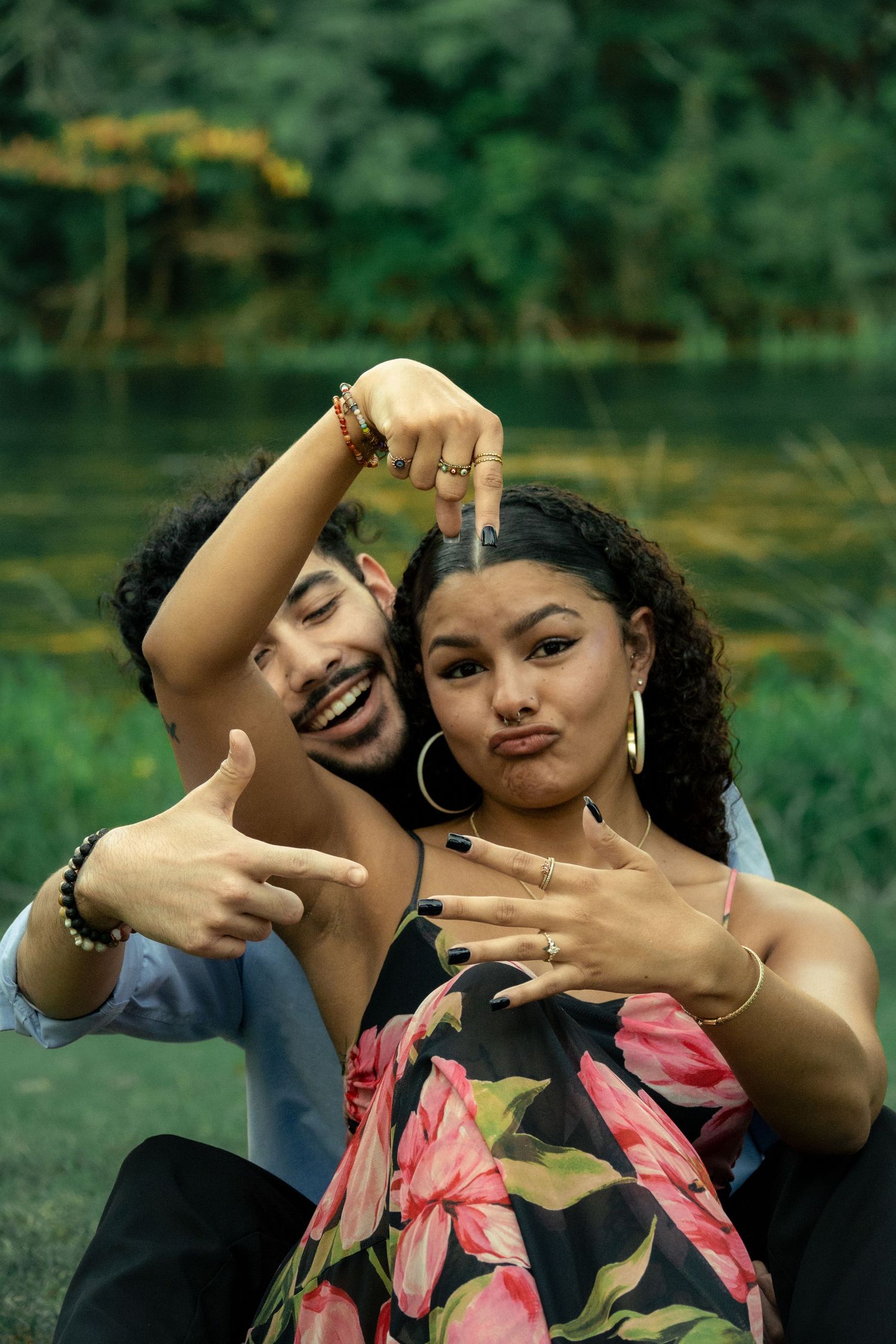 A young couple poses playfully outdoors near a pond, framing hands together, wearing colorful accessories and floral dress.
