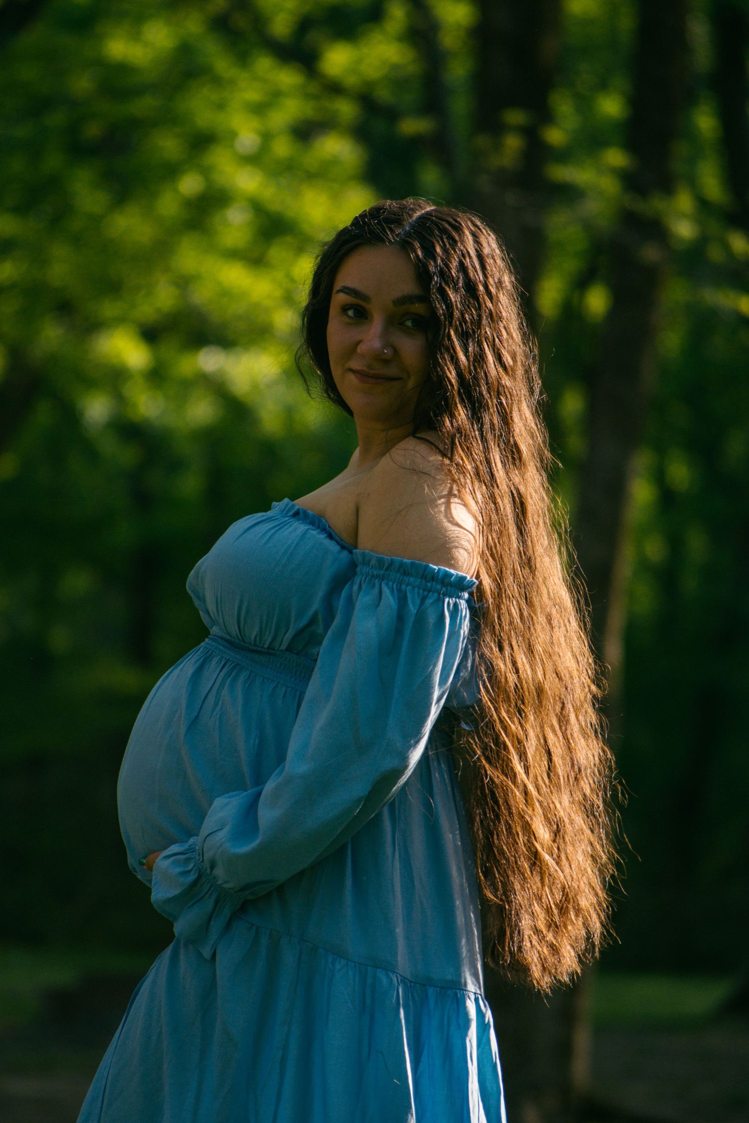 Pregnant woman in blue off-shoulder dress with long wavy hair posing in green forest.