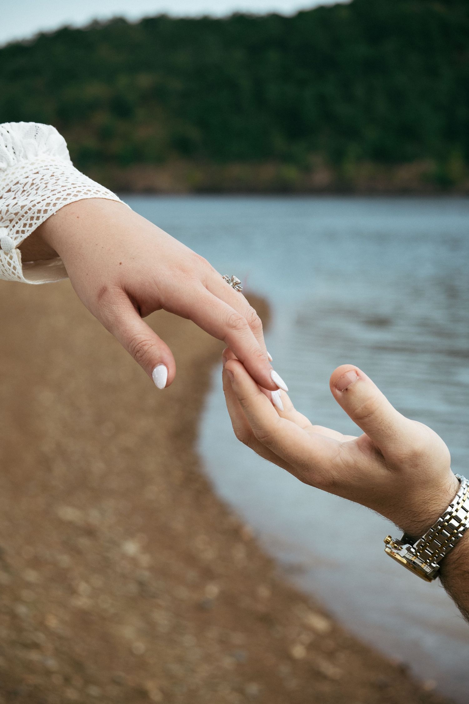 Two hands gently touching near a lakeside shore, one wearing a ring and lace sleeve, the other a gold watch.