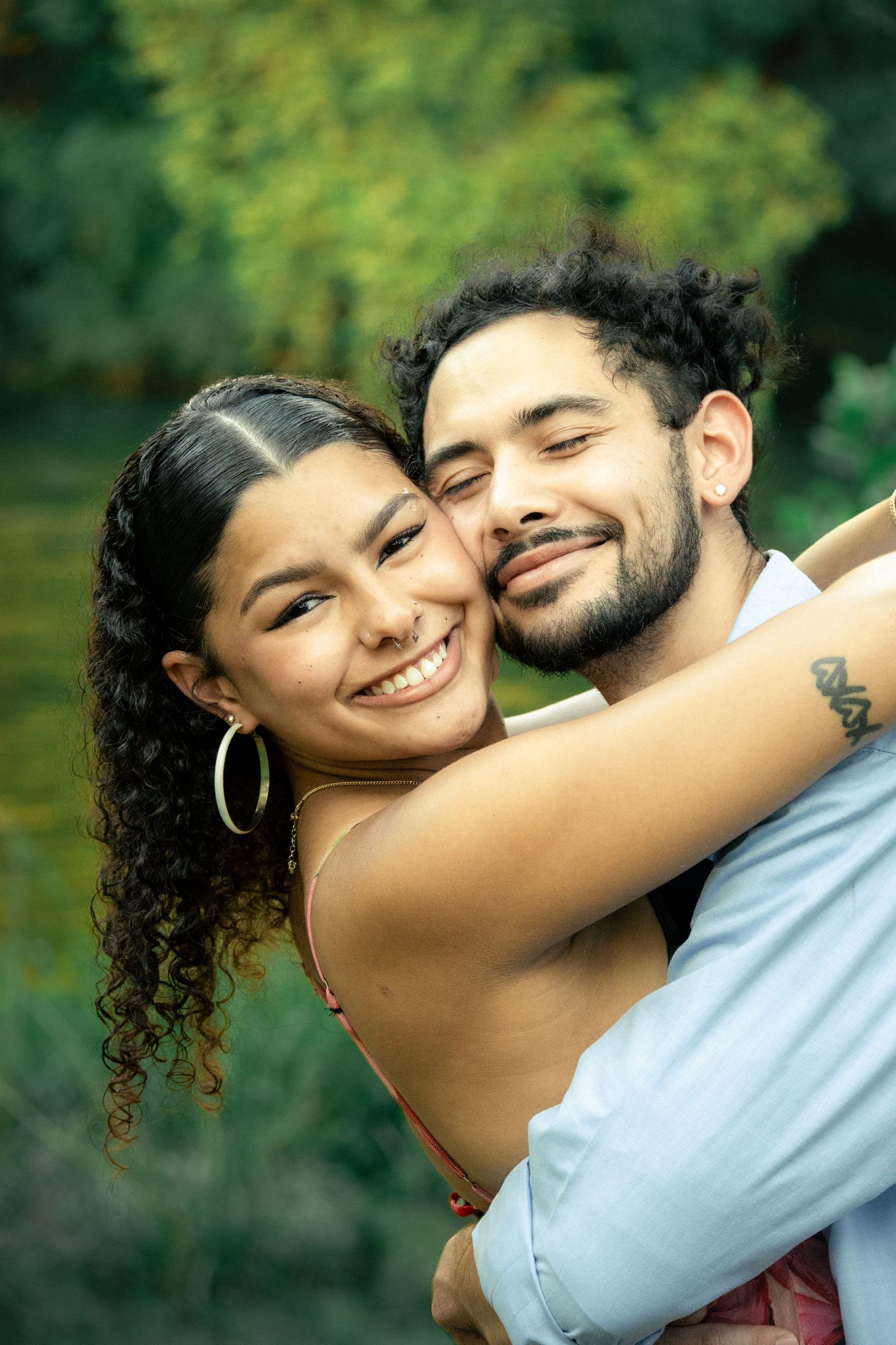 A smiling couple embraces outdoors, with a woman in hoop earrings and a man with a beard, surrounded by green foliage.