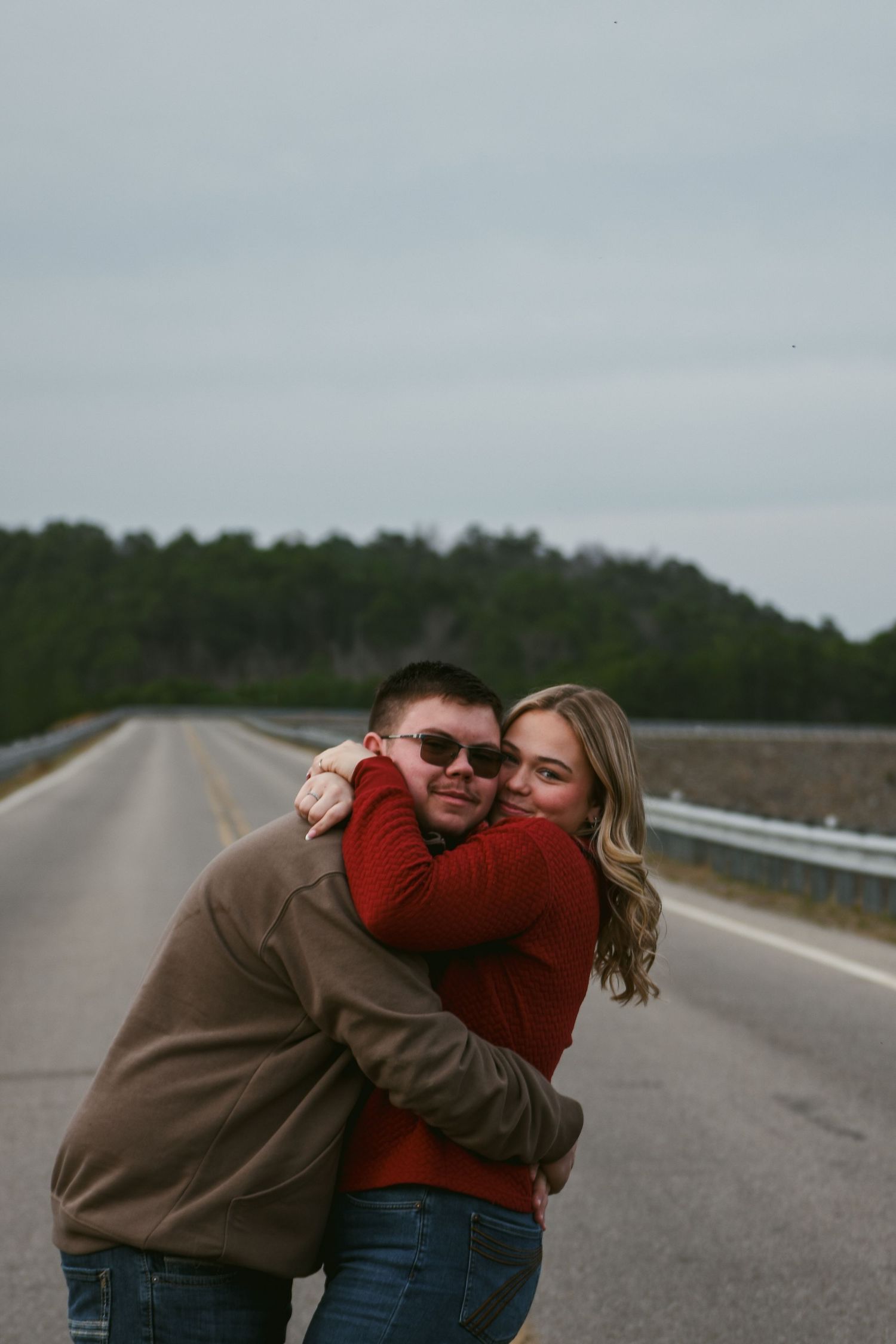 A smiling couple hugs on an empty road surrounded by pine trees under a cloudy sky.