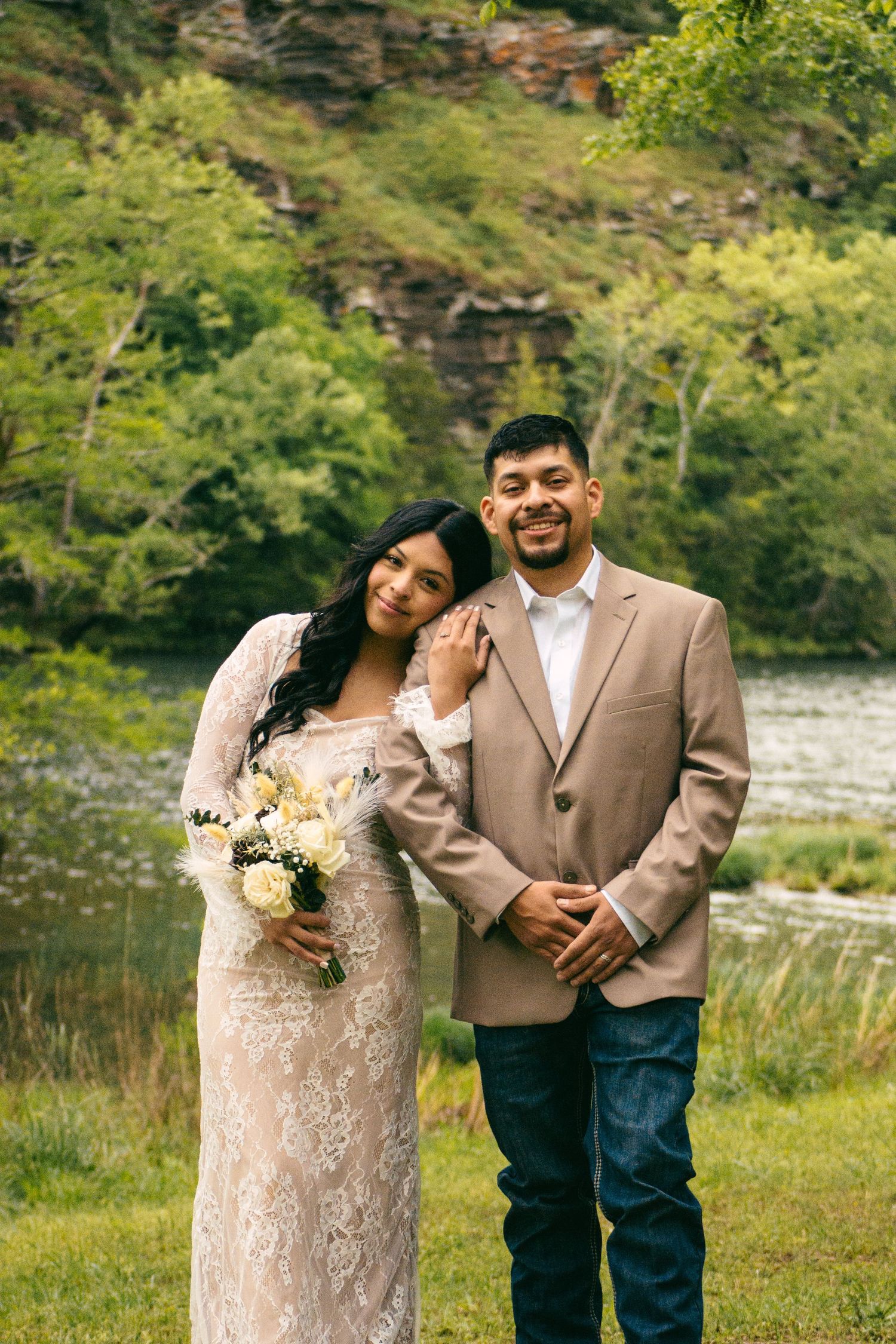 Bride in lace dress holding white bouquet leaning on groom in tan blazer by river.