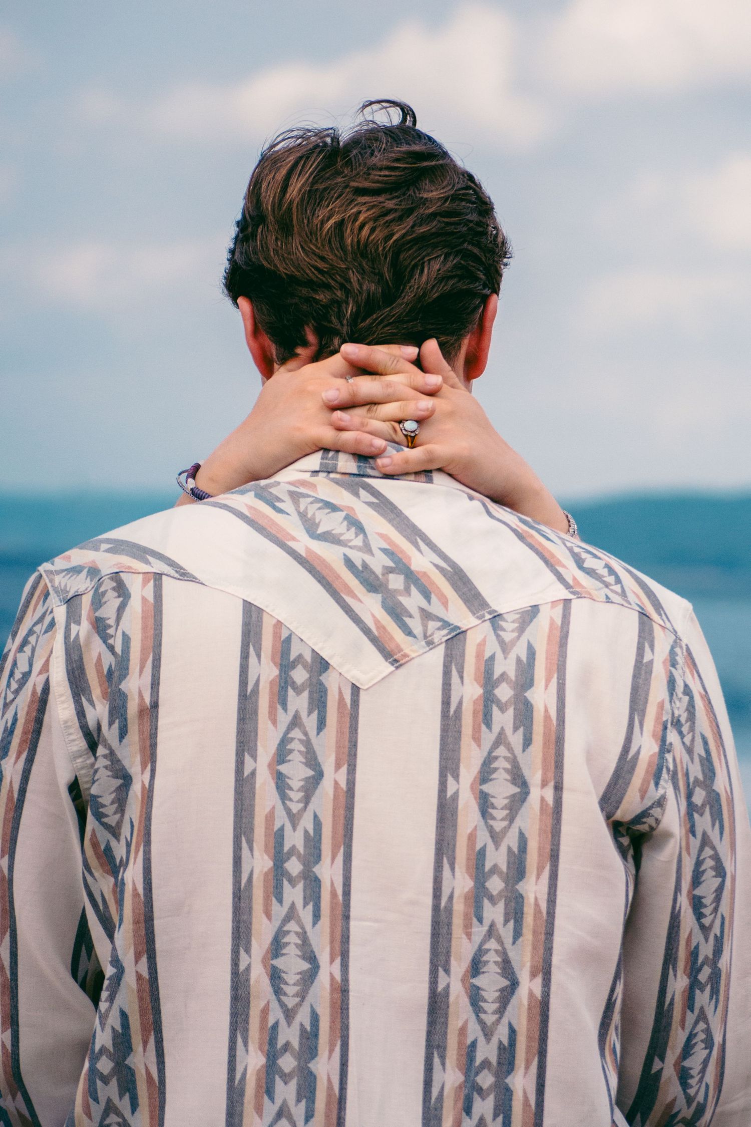 Back view of man in patterned Western shirt with hand resting on neck against blue sky and water.