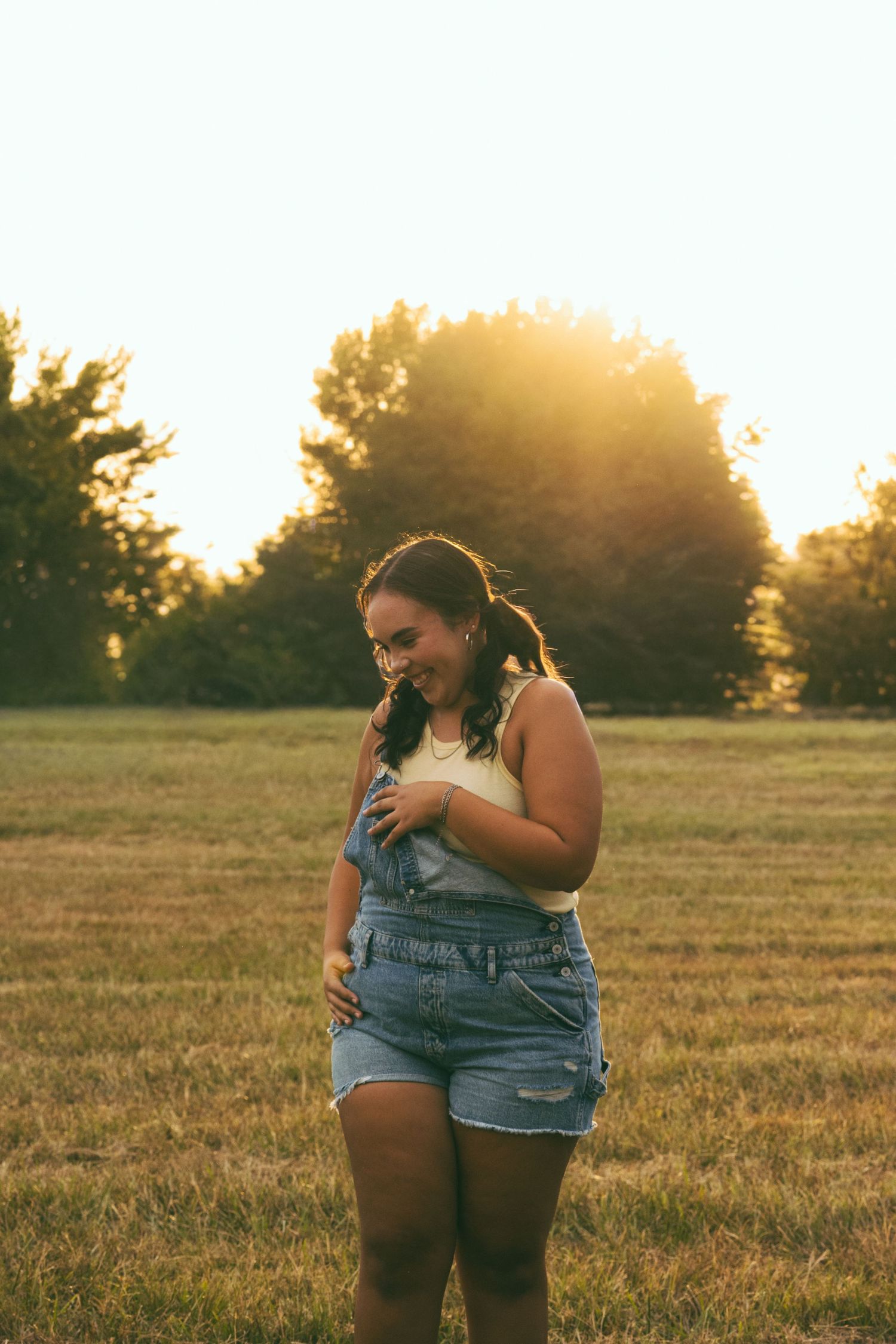 Young woman in denim overalls smiling in a golden sunlit field with trees in the background at sunset.
