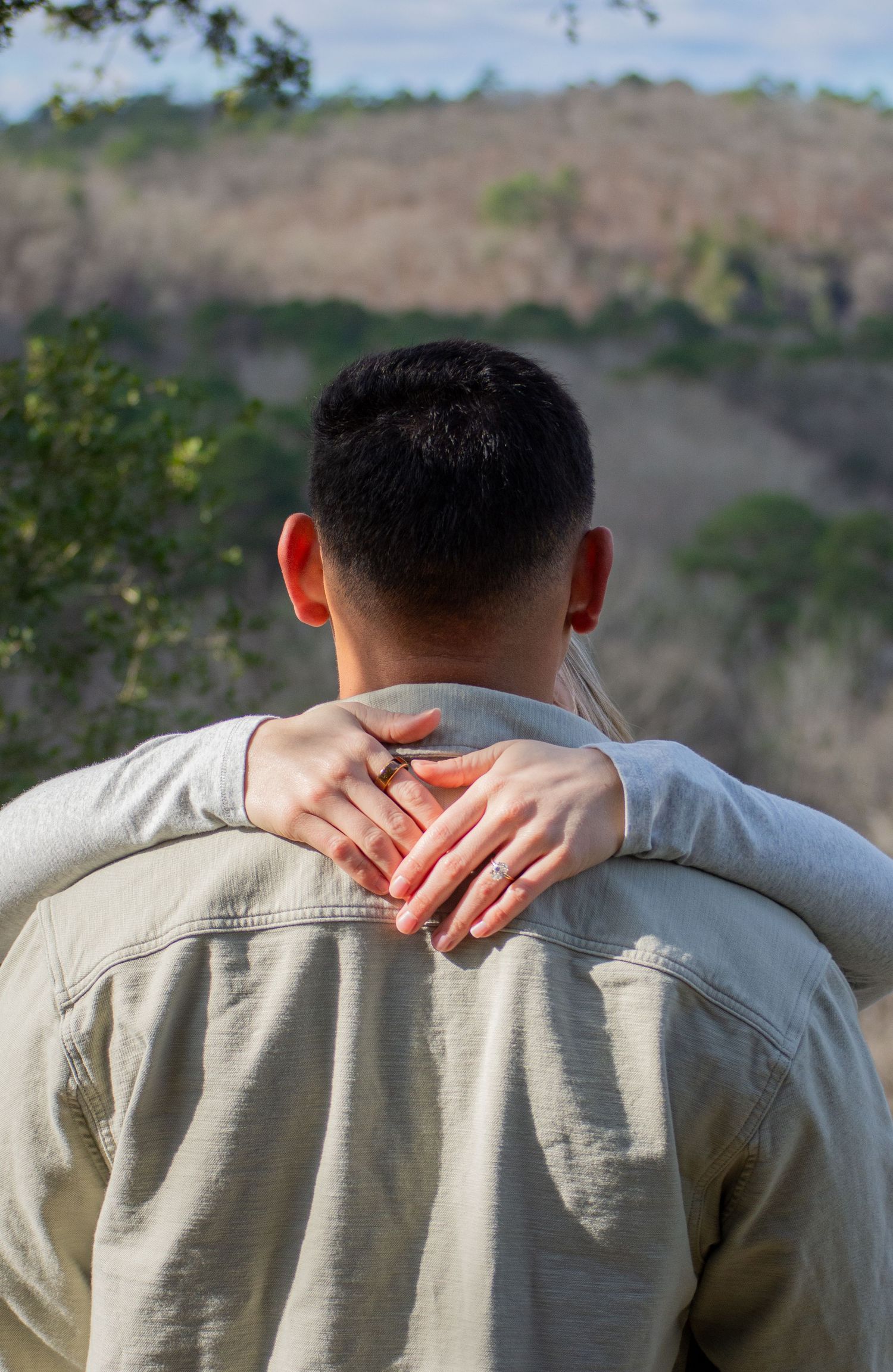 A woman's hand with a ring rests on a man's shoulder as they look out at a scenic landscape.
