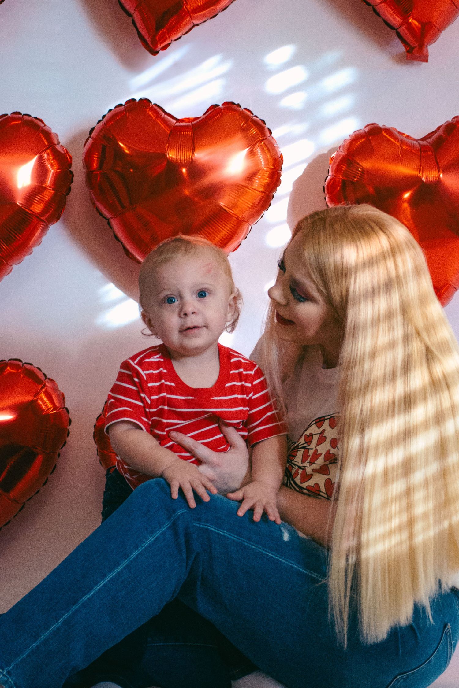 Blonde mother sitting with baby boy surrounded by red heart-shaped balloons on Valentine's Day.