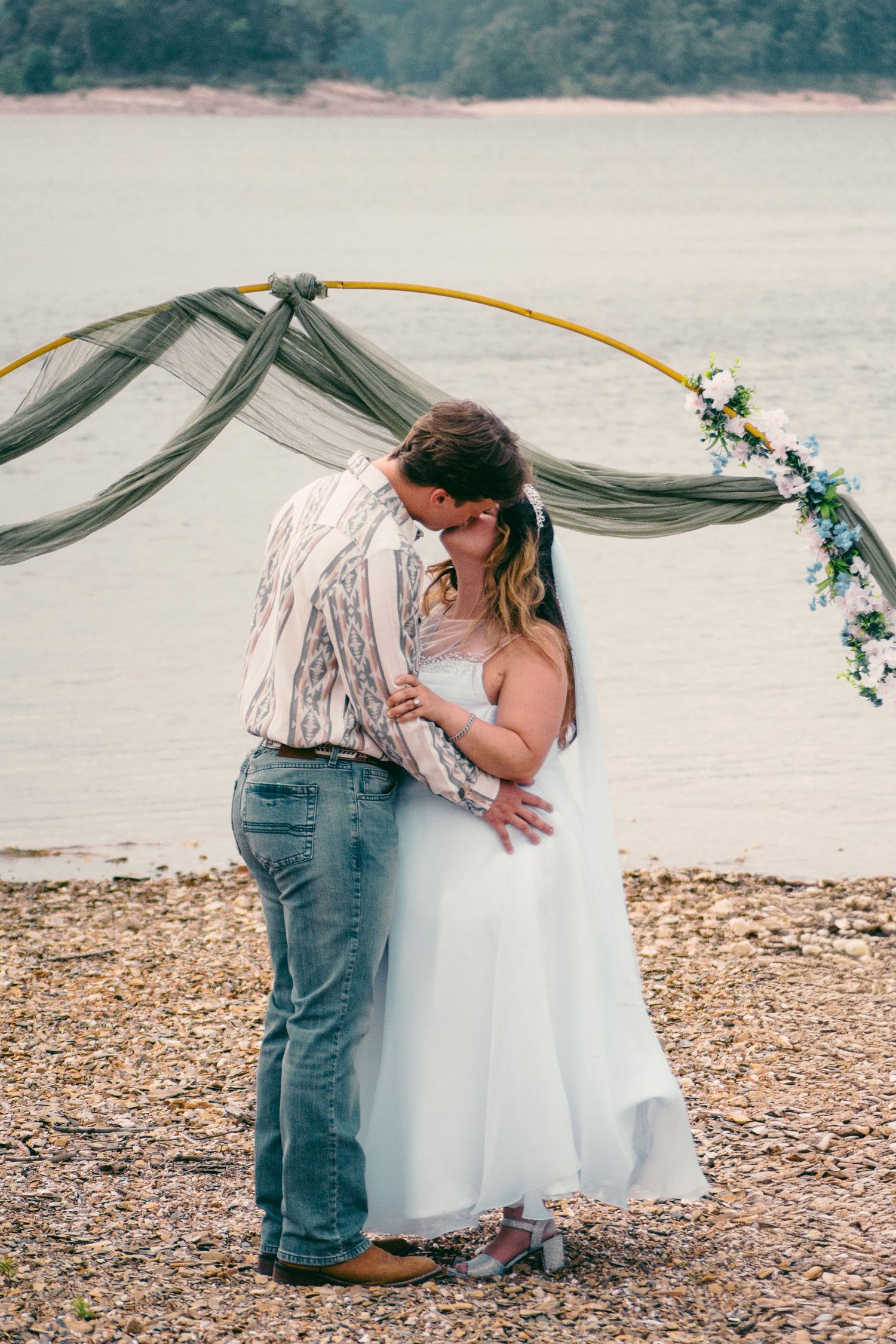 A bride and groom share a first kiss under a floral hoop arch on a rocky lakeside shore.