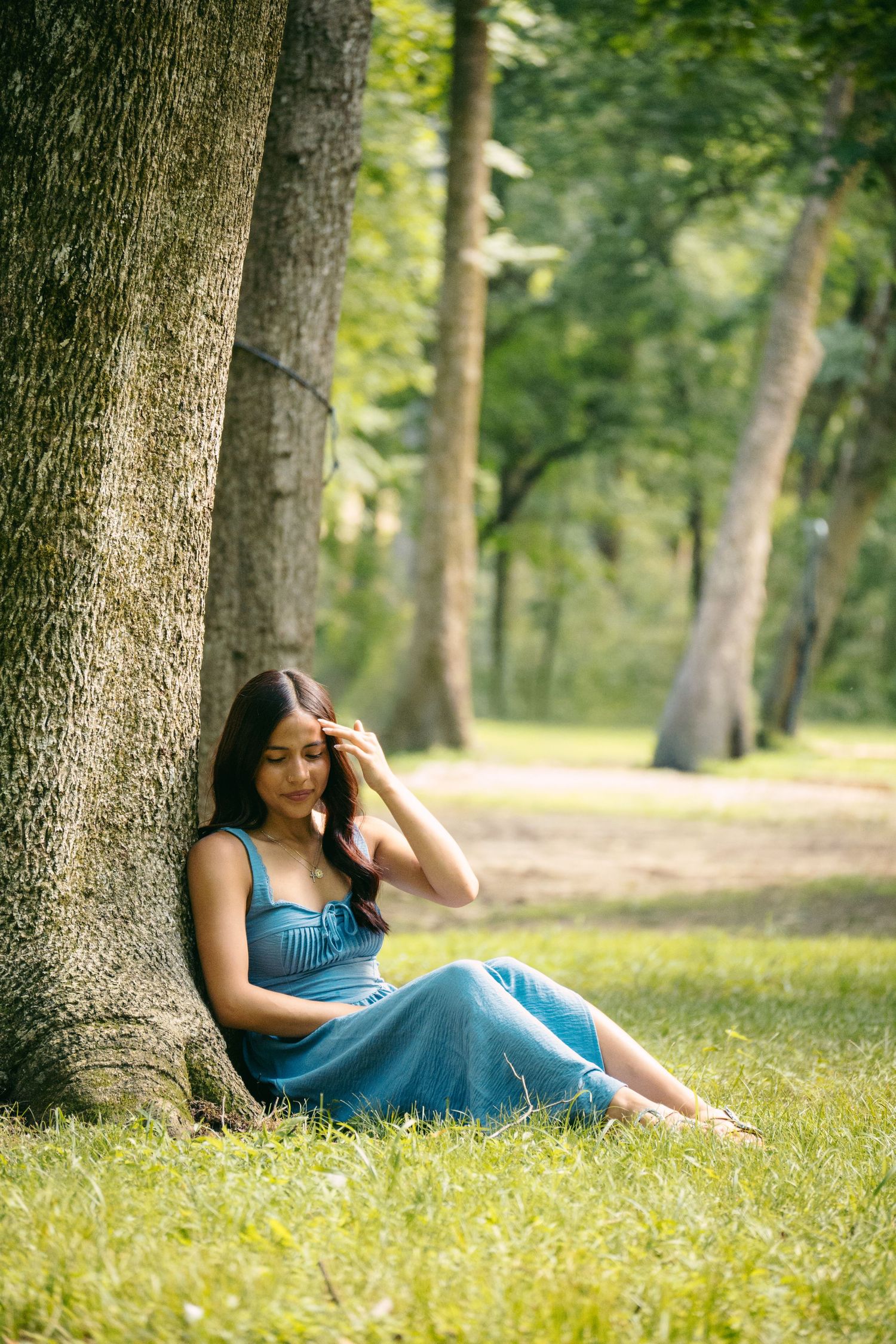 Young woman in a blue dress sitting against a tree trunk on green grass in a sunny park.