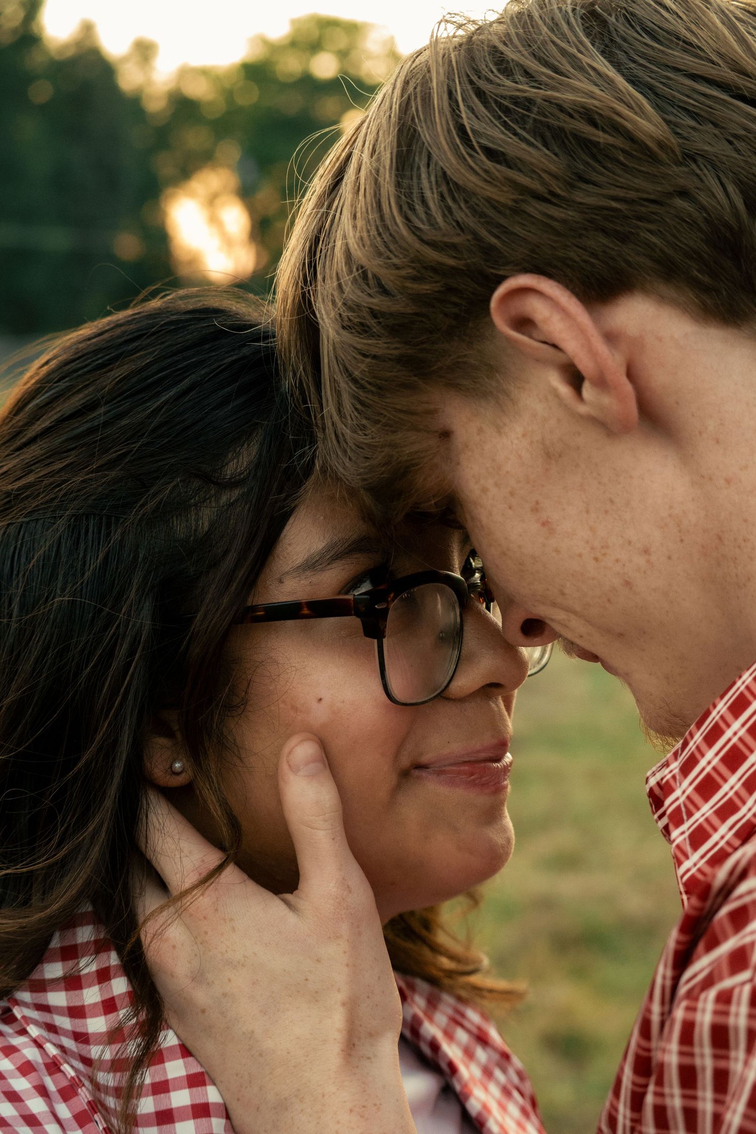 A couple shares an intimate moment outdoors at golden hour, foreheads touching, woman wearing glasses and red plaid shirt.