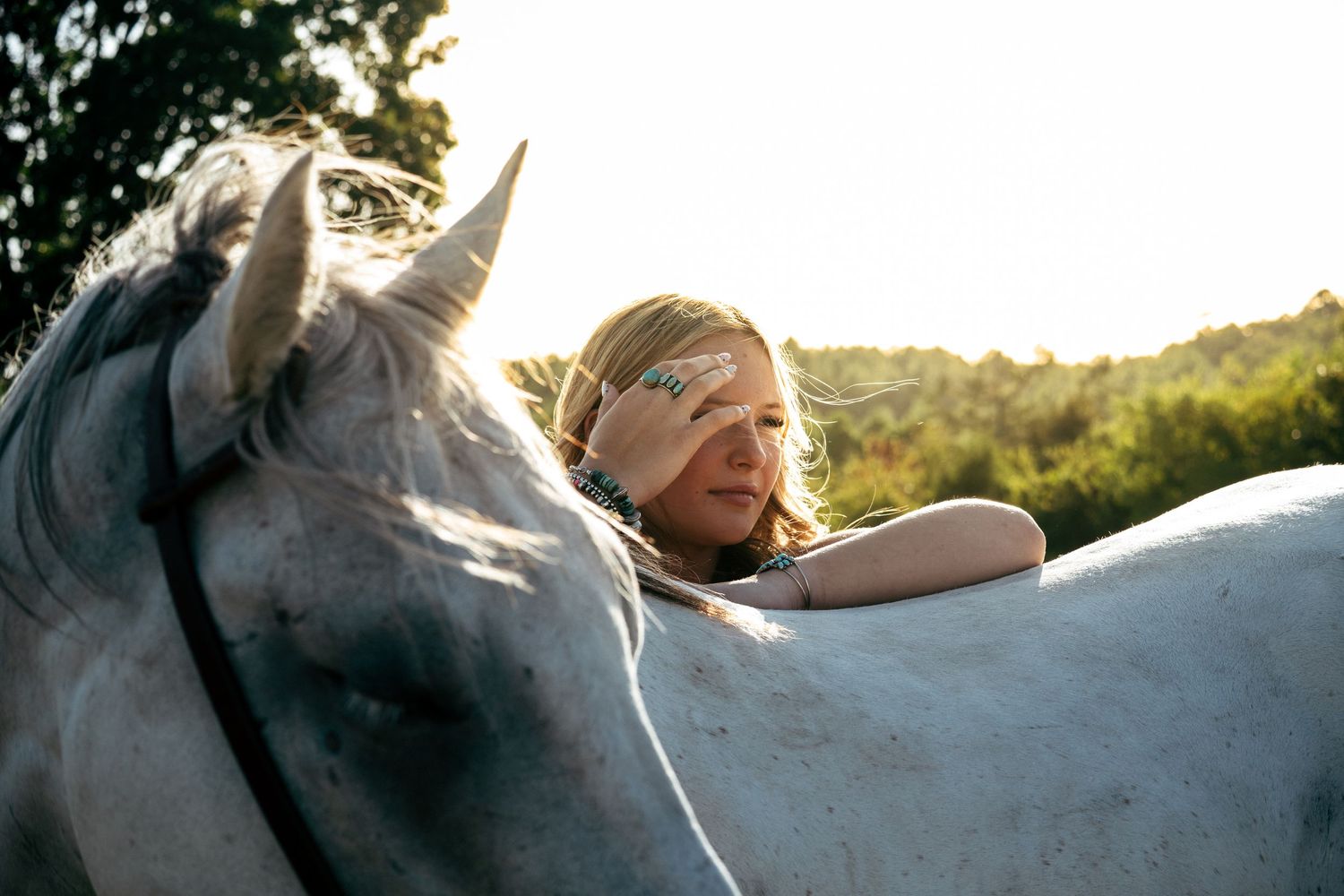Young blonde woman resting her head on a white horse outdoors at golden hour.