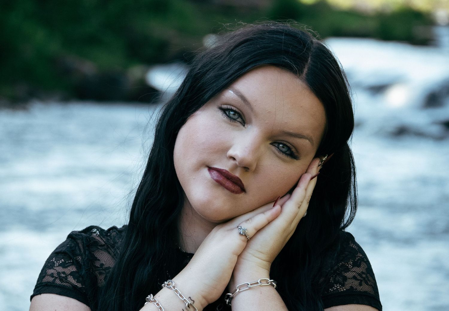Young woman with dark hair posing gracefully near a river, wearing a black lace top and delicate bracelets.