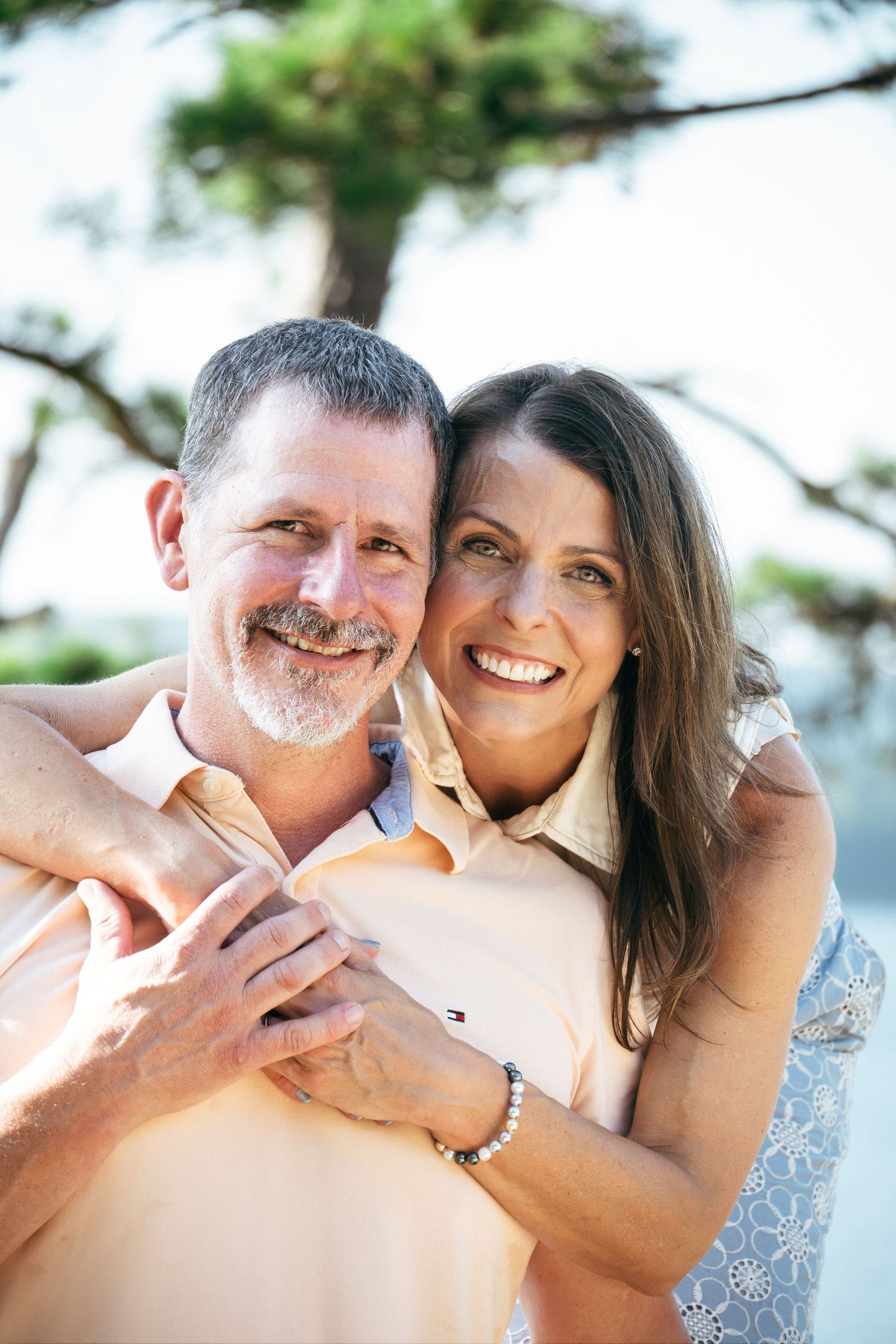 Happy middle-aged couple embracing outdoors, smiling warmly near a tree on a sunny day.