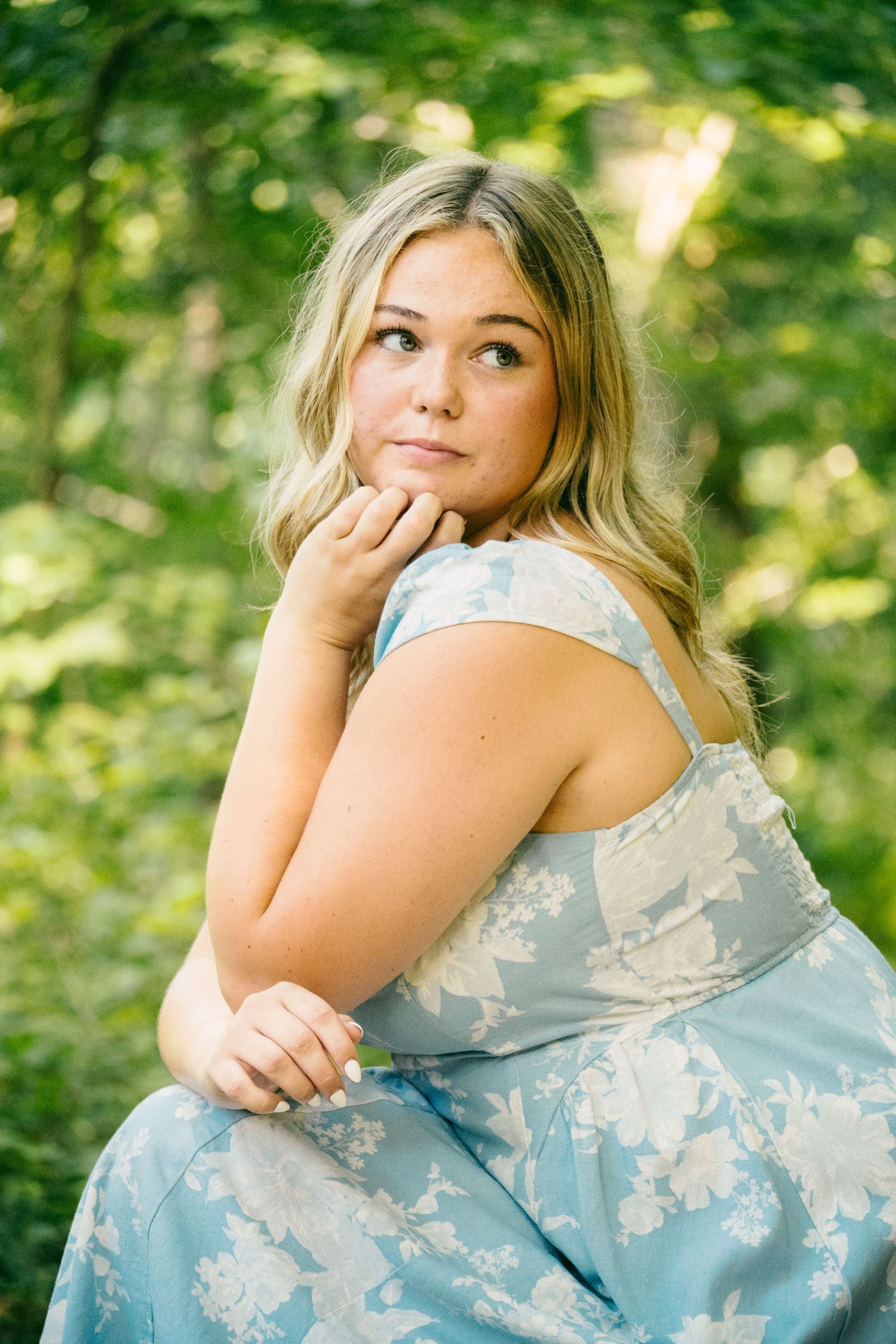 Young blonde woman in a floral blue dress posing thoughtfully outdoors in a lush green forest.