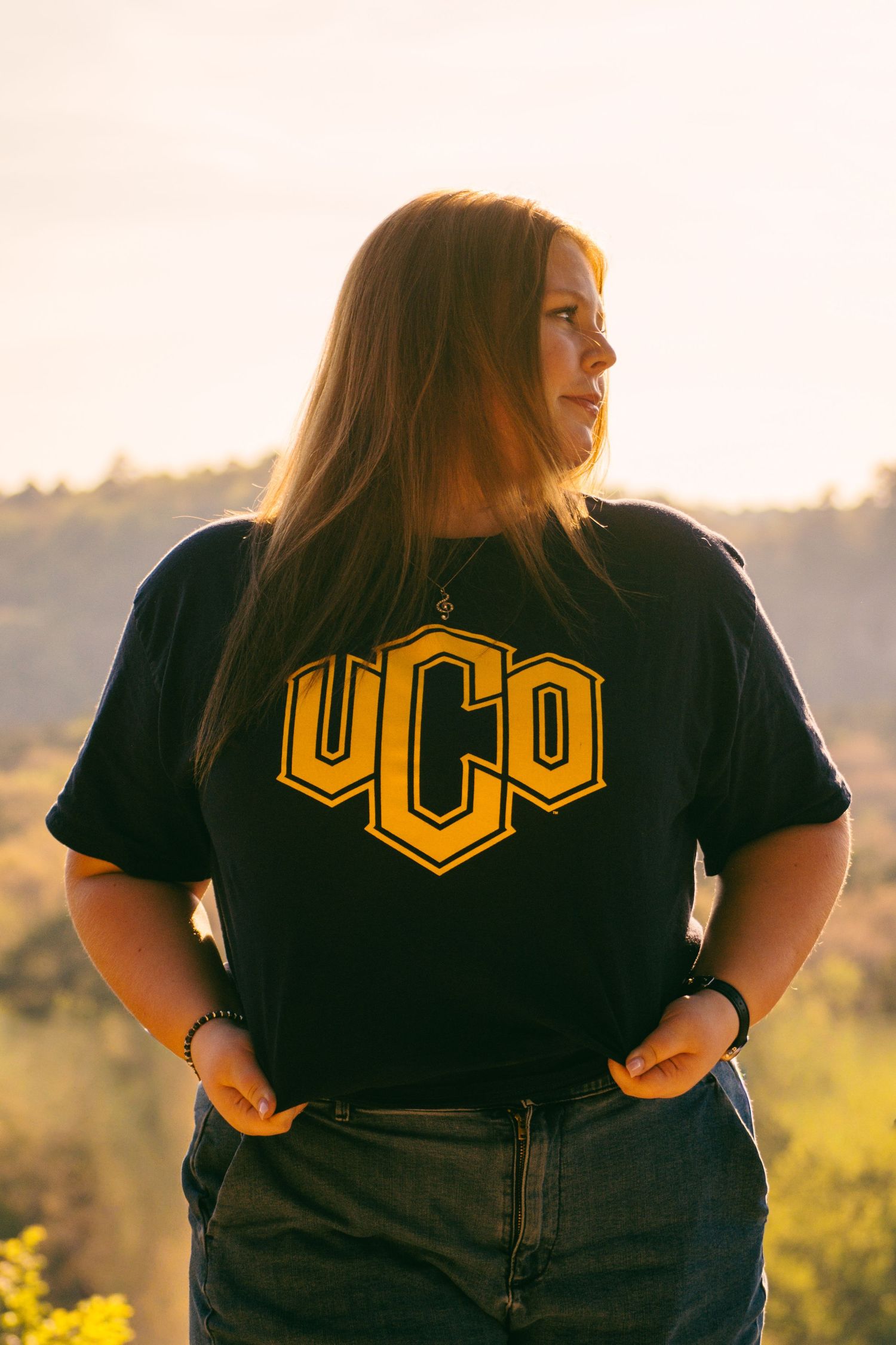Young woman wearing a UCO black and gold t-shirt standing outdoors at golden hour with scenic hills behind her.
