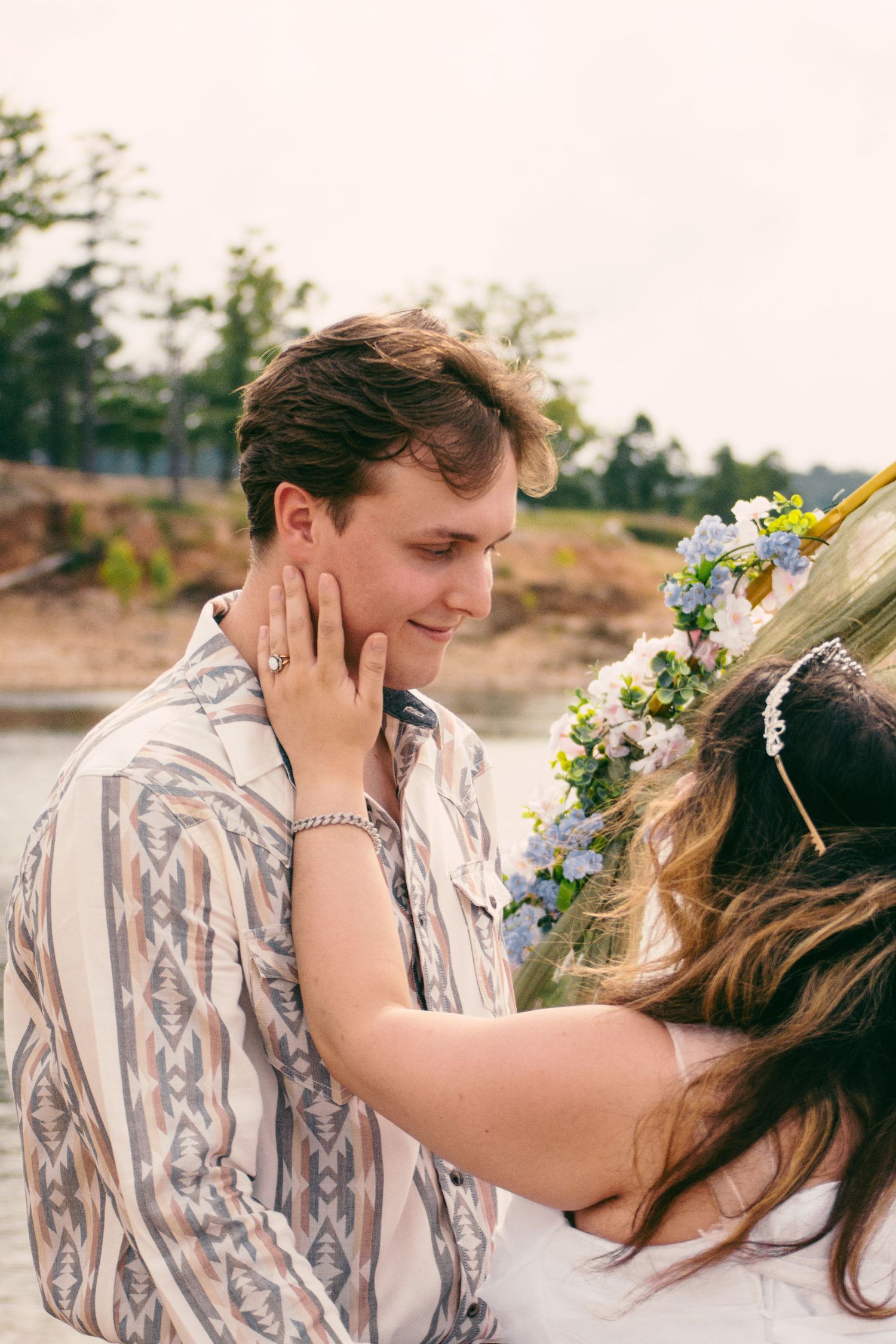 Bride wearing floral crown gently touching groom's face during romantic outdoor wedding moment.