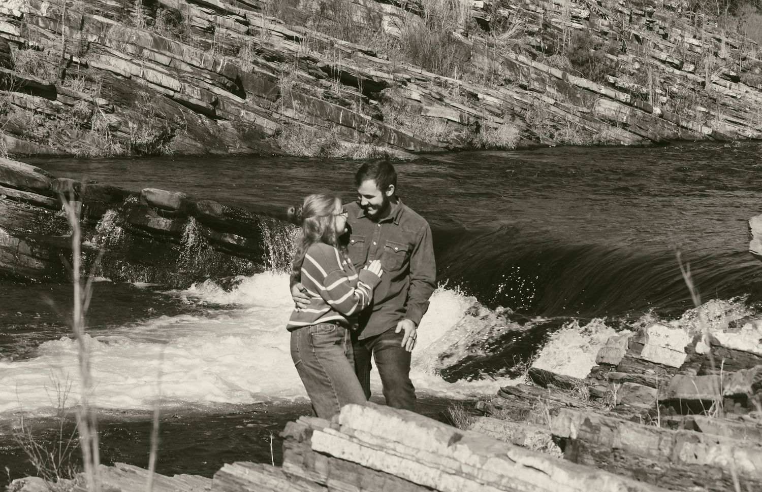 Couple embracing on rocks beside a rushing waterfall with layered stone cliffs in the background.