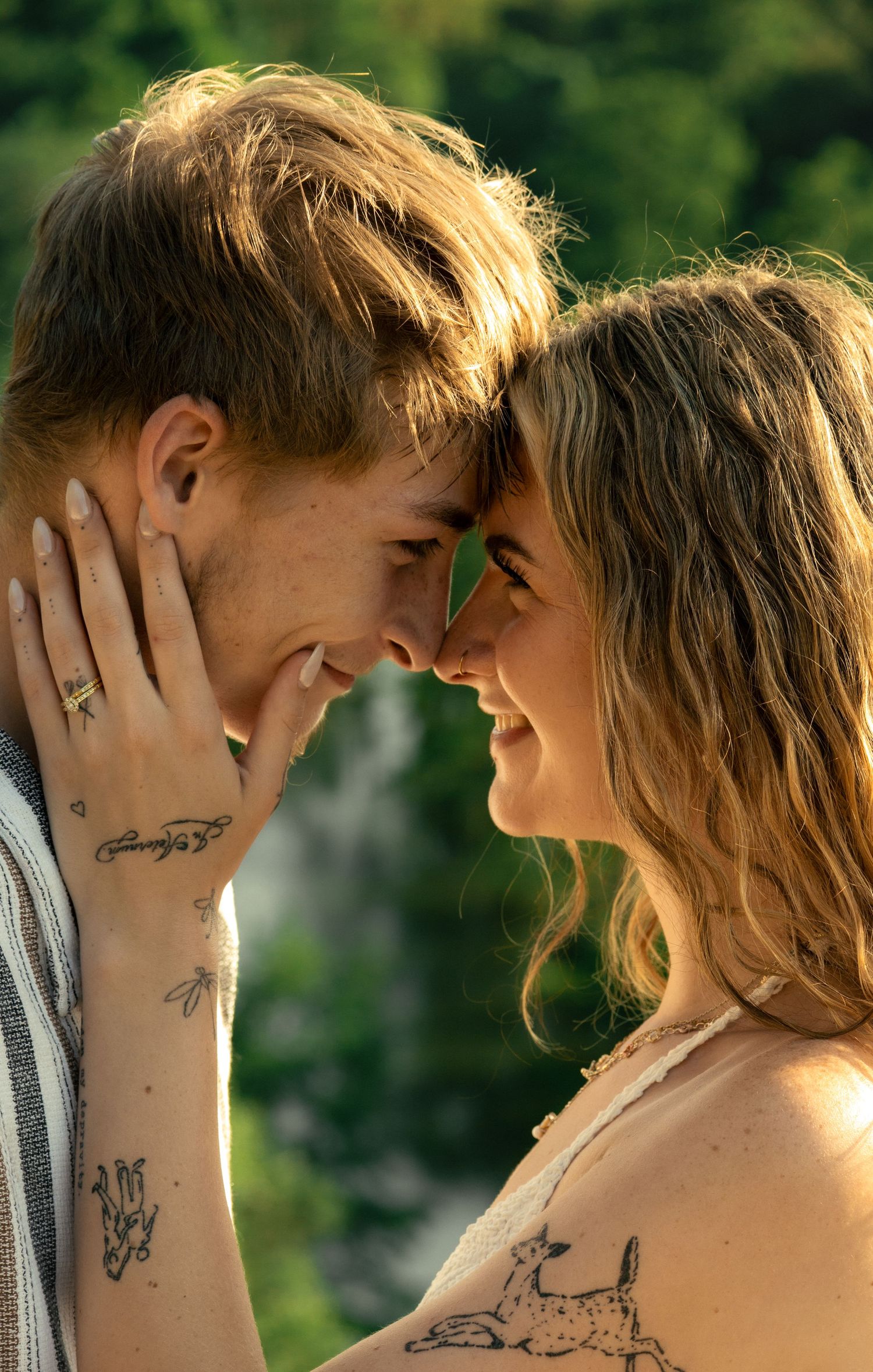A couple with tattoos sharing a romantic forehead touch, smiling in warm golden sunlight with green foliage behind them.