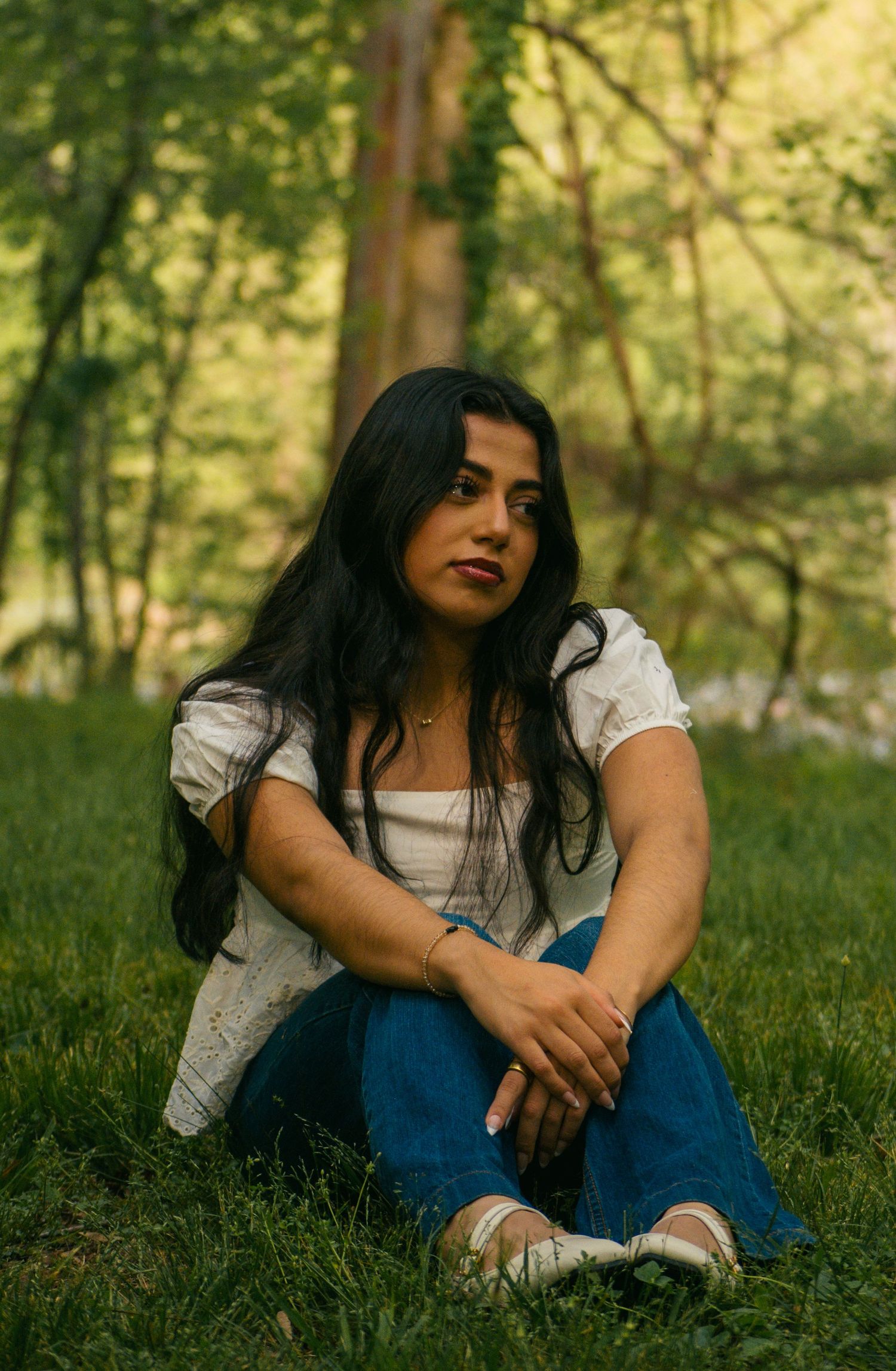 Young woman with long dark hair sits on grass in a lush green forest, wearing white top and blue jeans.