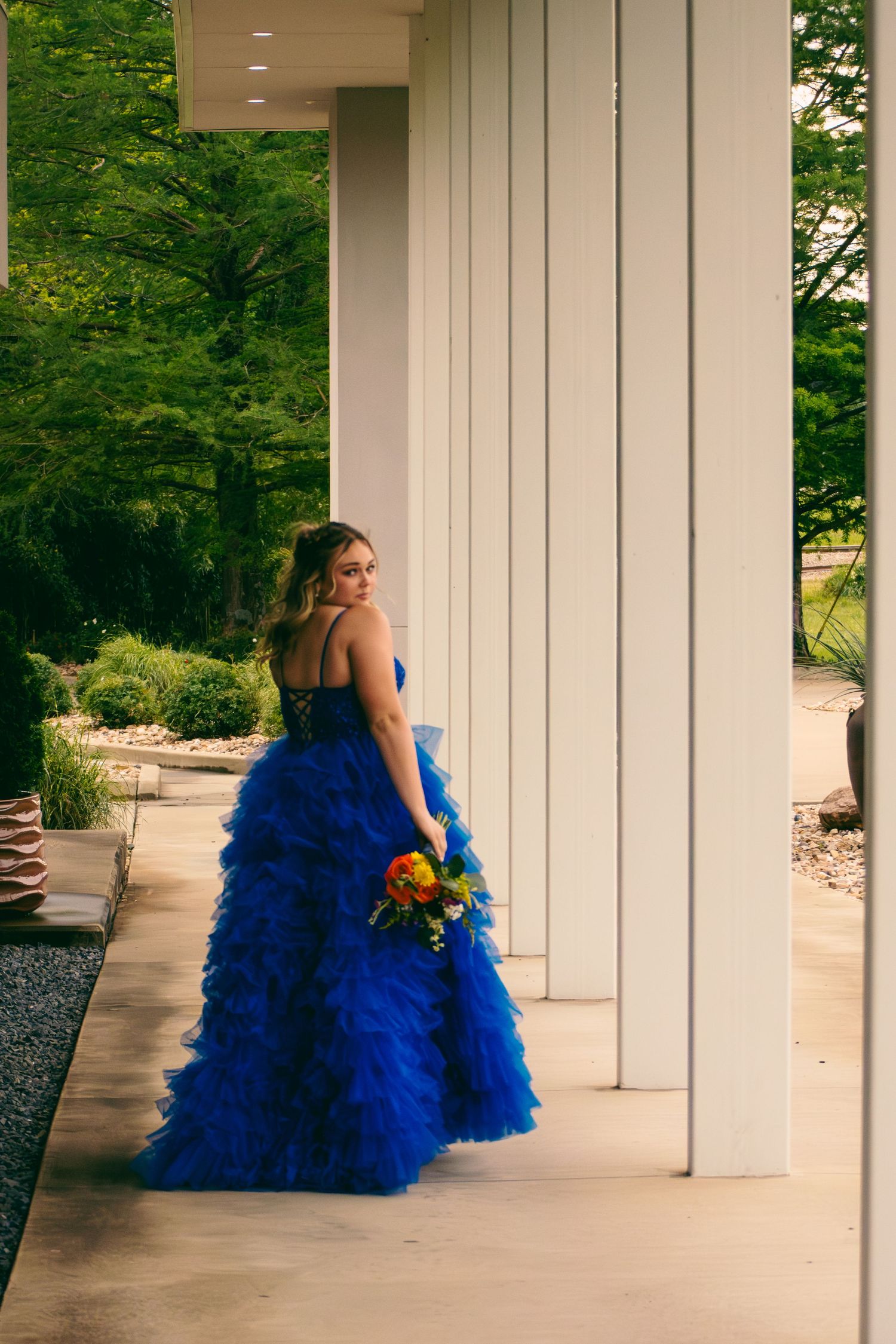 Young woman in stunning royal blue ruffled prom dress holds colorful bouquet between white columns outdoors.