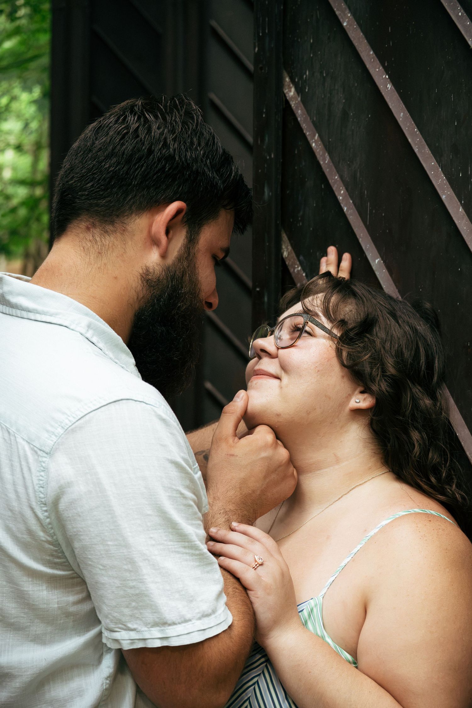 A bearded man tenderly holds a smiling woman's chin against a dark wooden wall outdoors.